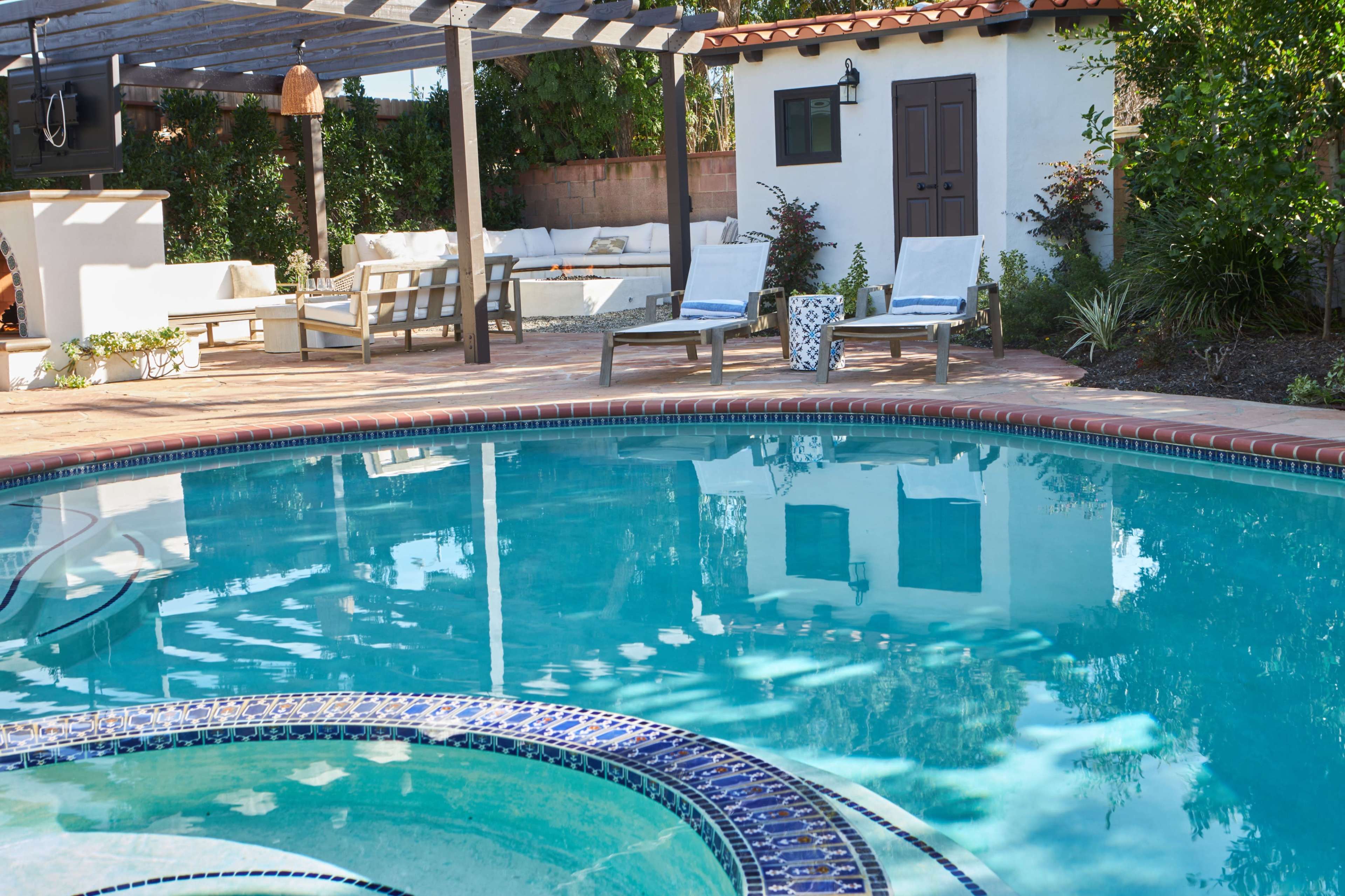 The image shows a sunny backyard with a swimming pool, loungers, and a covered seating area beside a white-walled building.