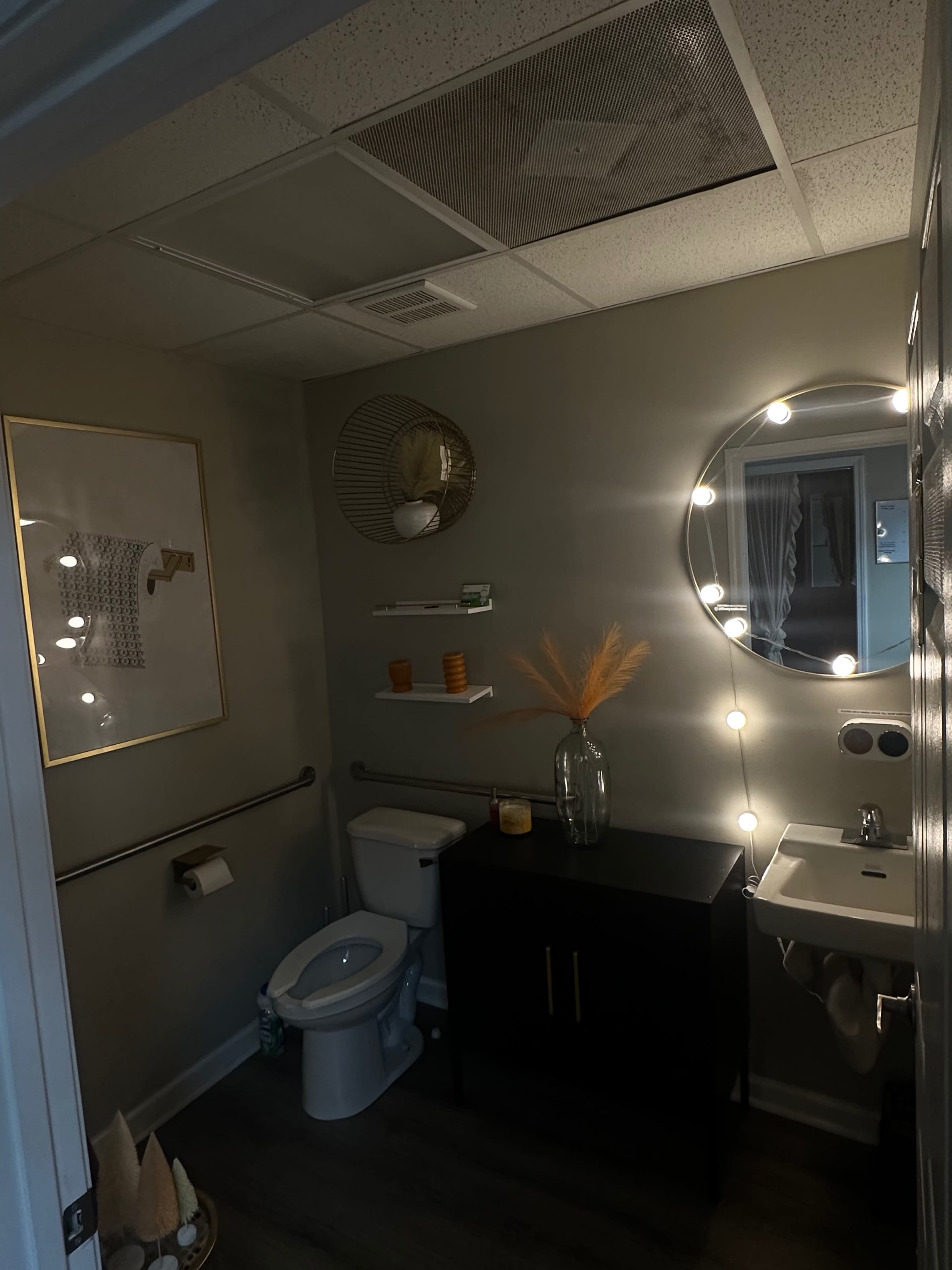 A modern bathroom features a toilet, a sink, a decorative mirror with lights, and minimalist decor including a vase and wall art.