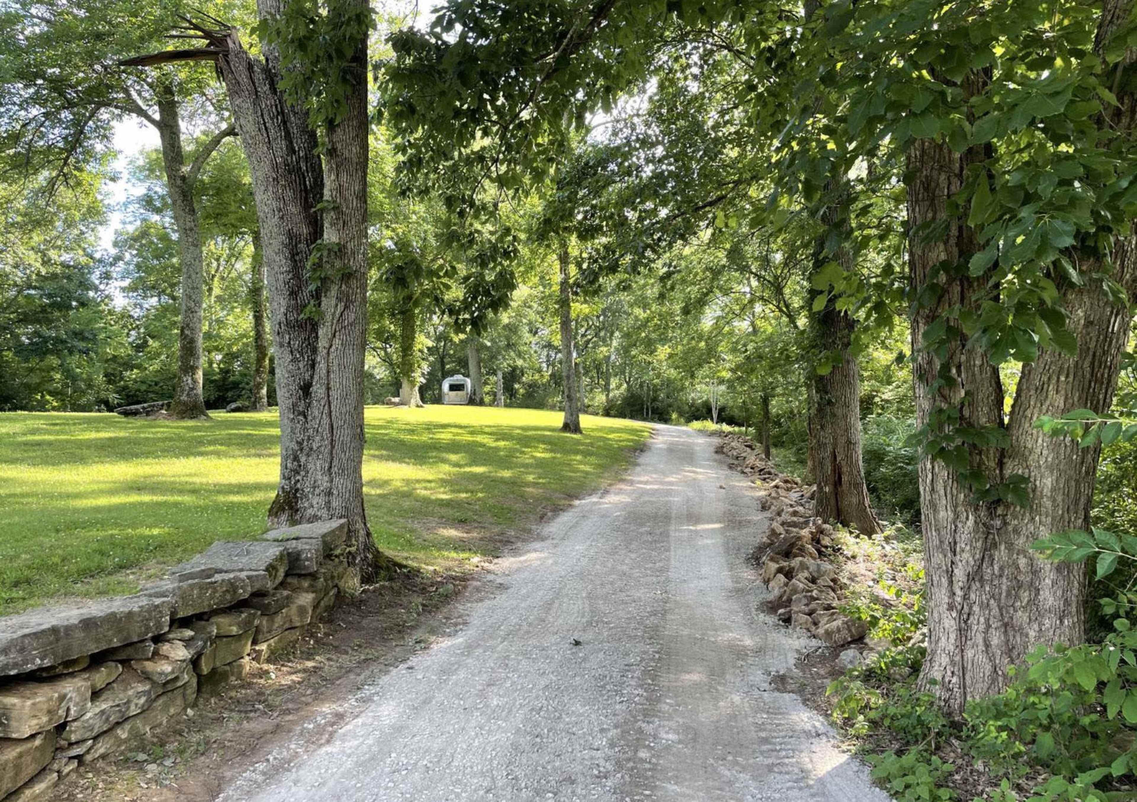 A gravel path winds through a tree-lined area with a grassy slope and stone border.