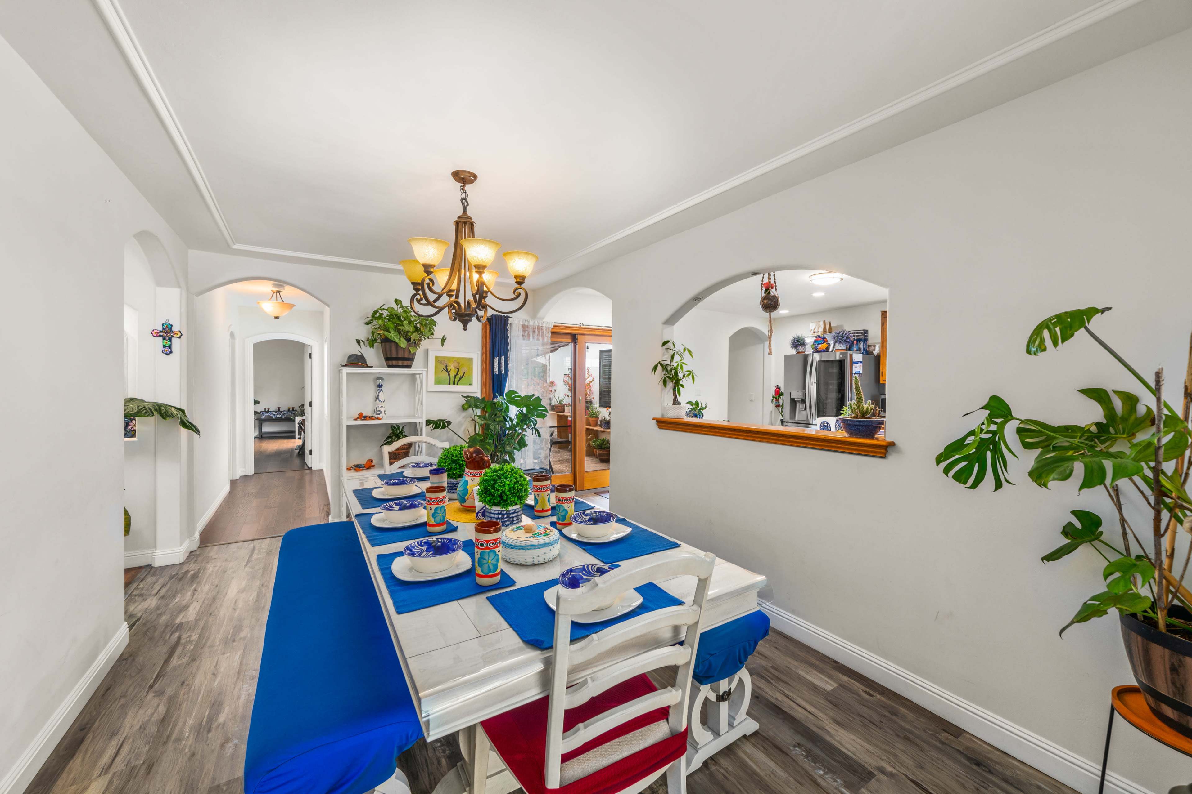 The image shows a dining area with a table set for a meal, featuring colorful table settings and surrounded by plants.