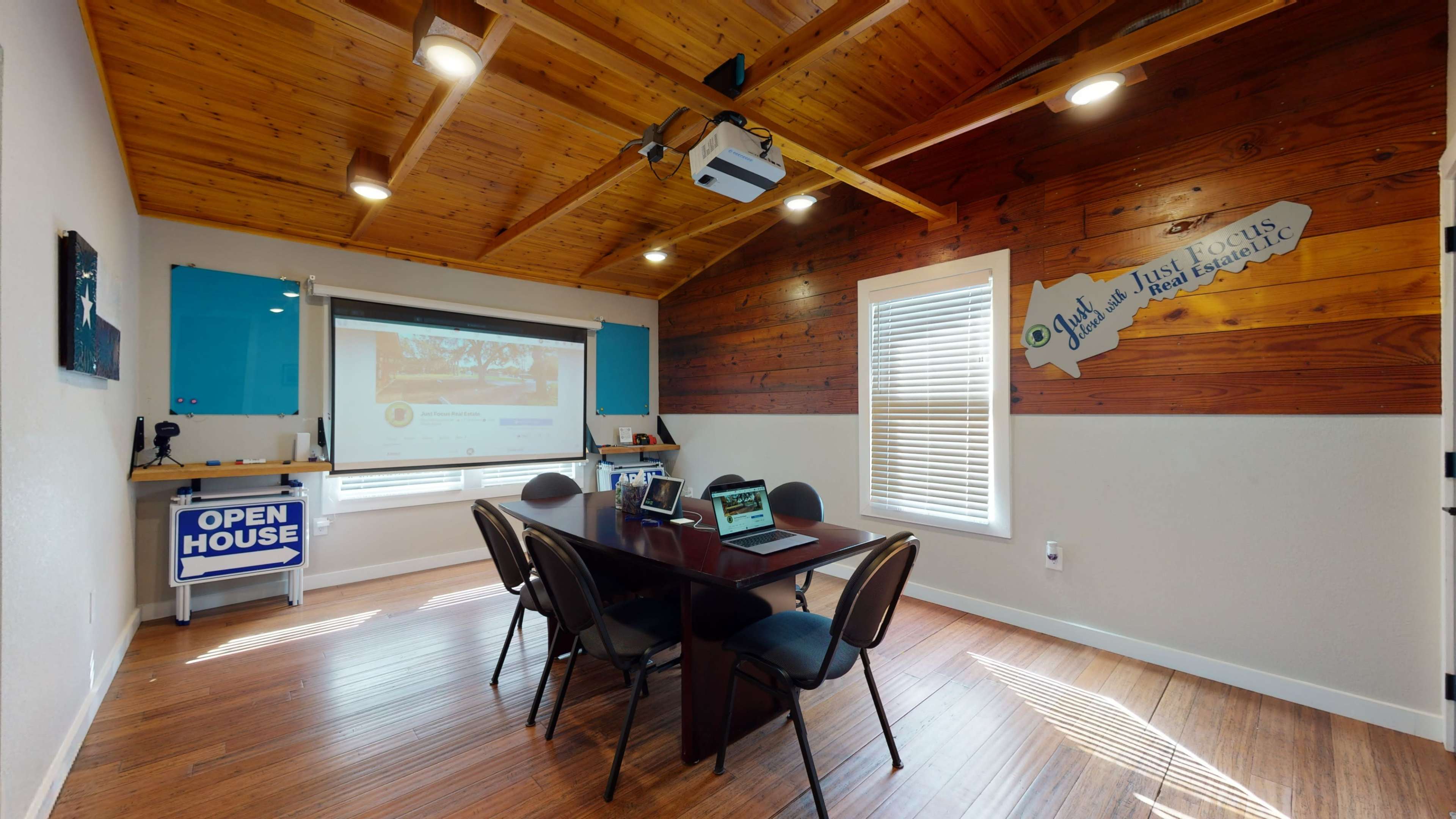 The image shows a modern conference room with a wooden ceiling, a large table surrounded by chairs, a projector screen on one wall, and various electronic devices on the table.