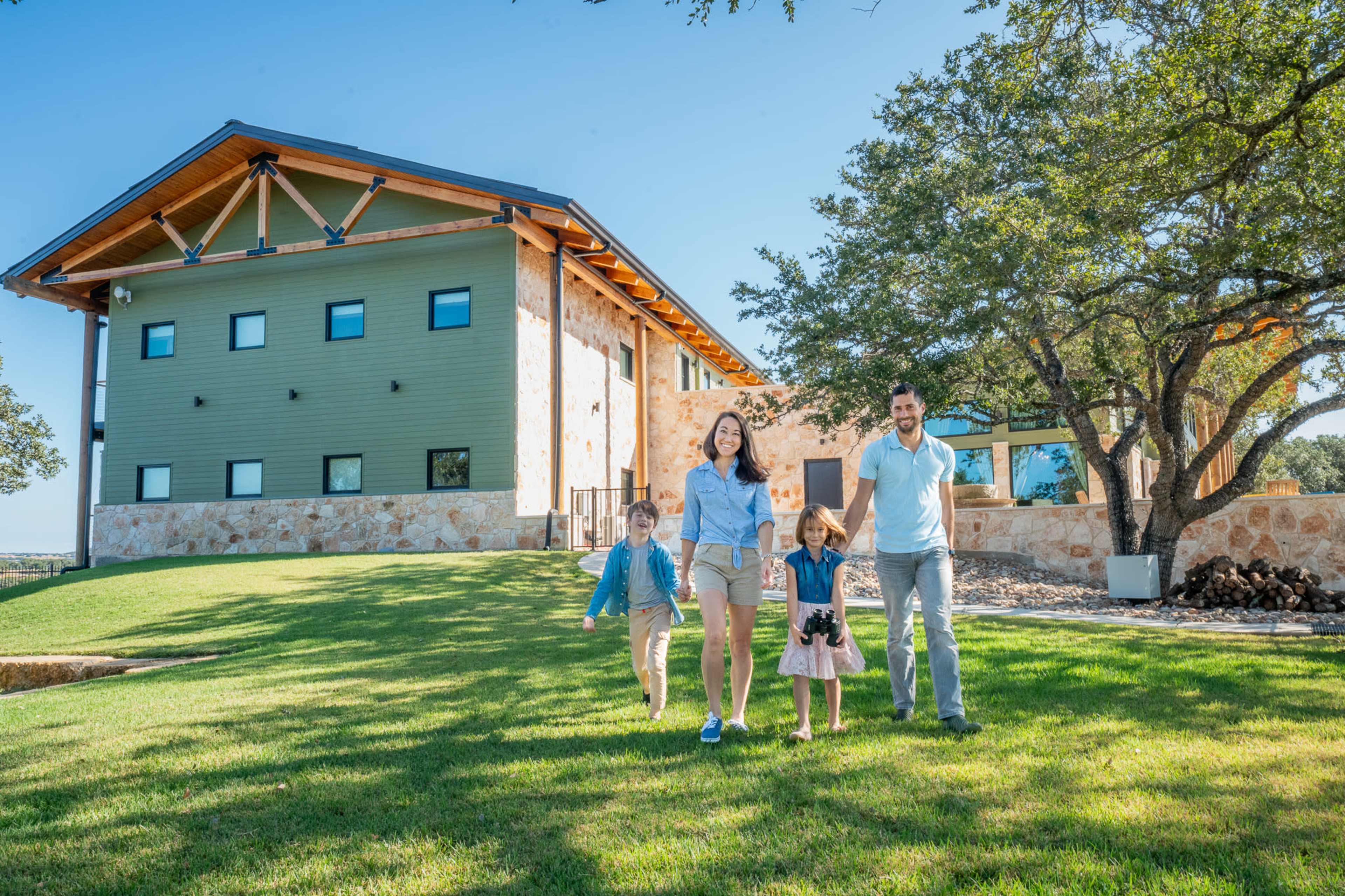 A family of four, including two children, walks together across a green lawn in front of a modern house with a stone exterior and a wooden roof.