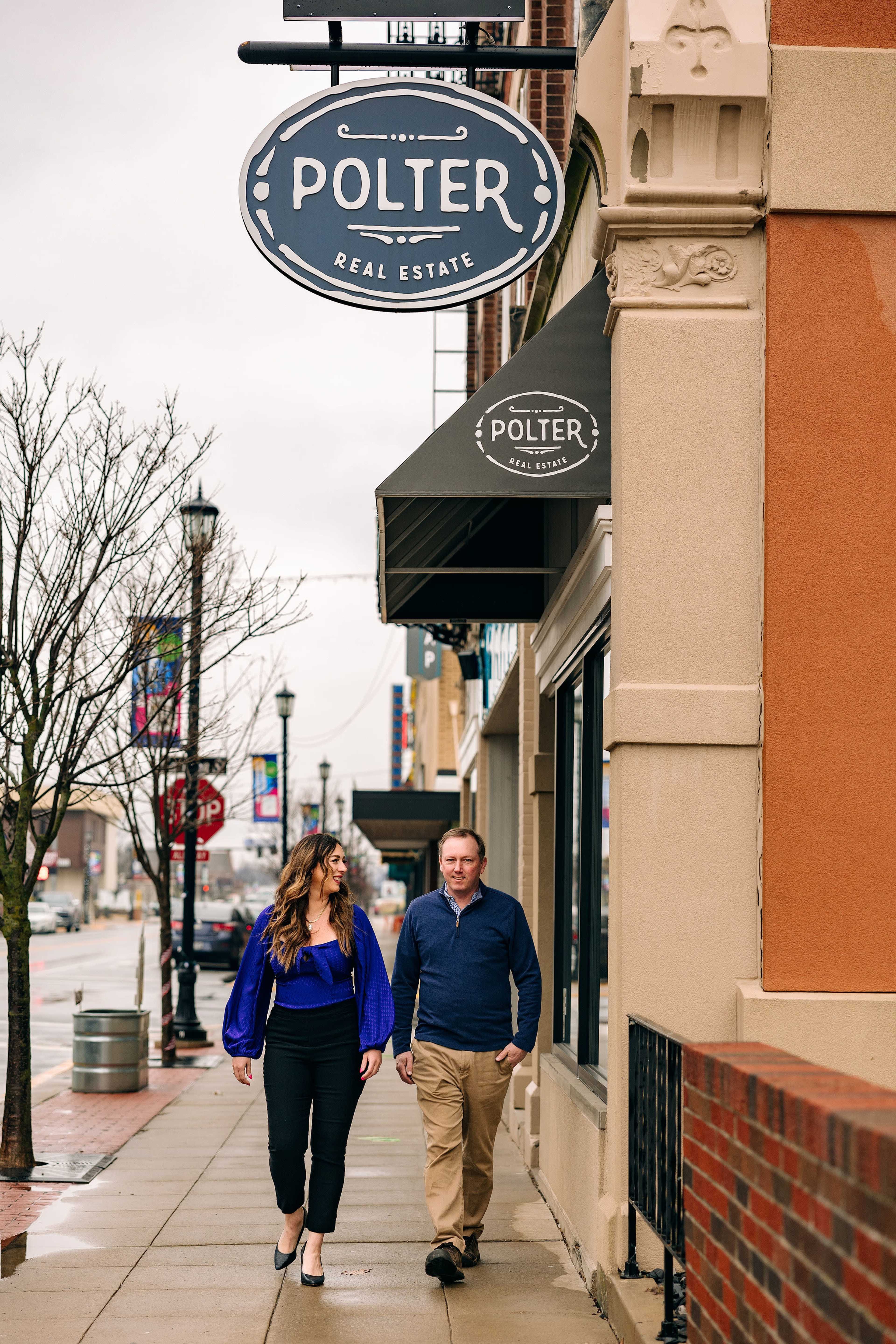 A woman and a man walk together along a sidewalk in front of a real estate office displaying a sign that reads "Polter Real Estate."