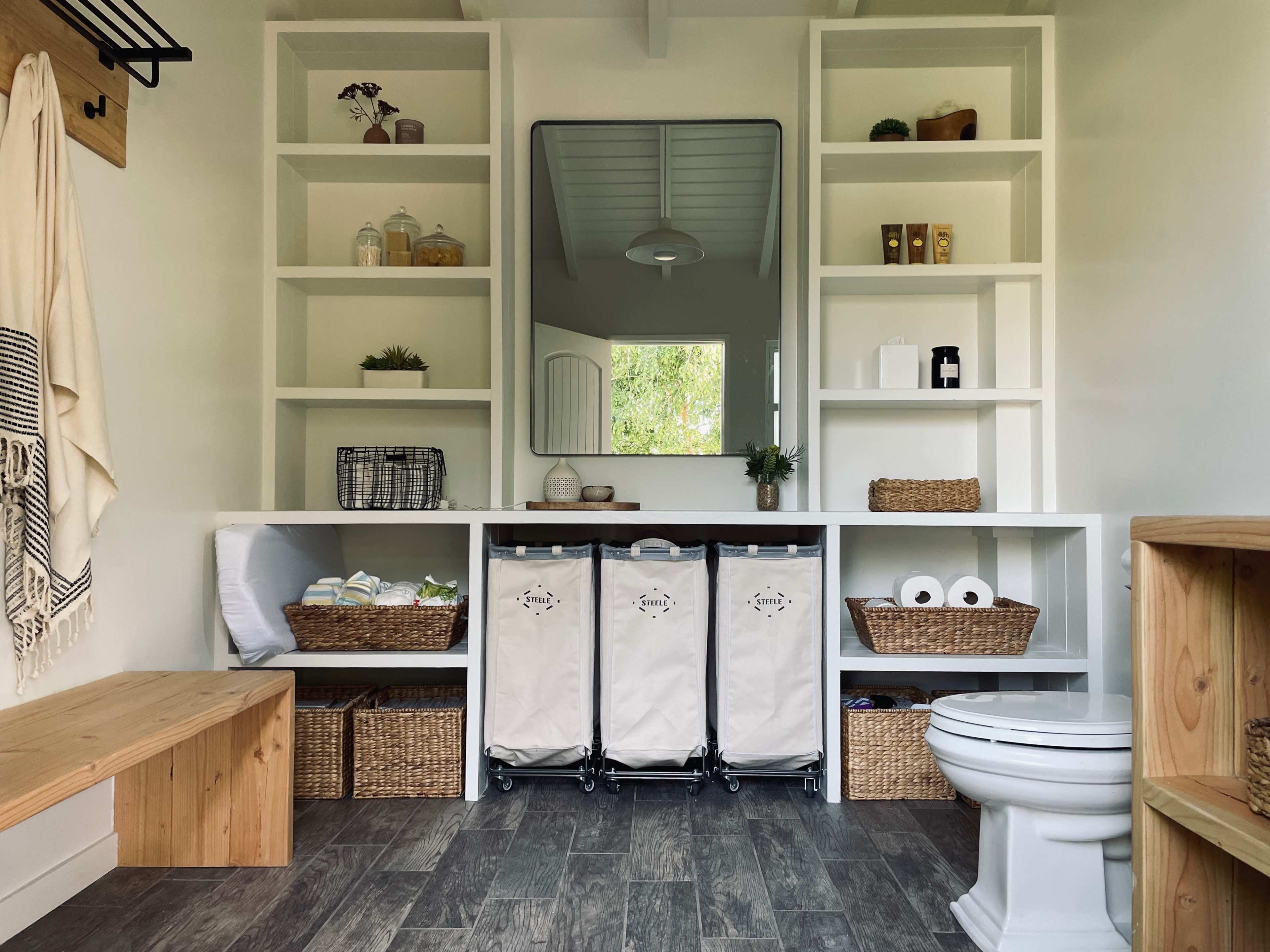 The image shows a minimalist bathroom with open shelving, a large mirror, and laundry bins, accompanied by wooden and wicker storage solutions.