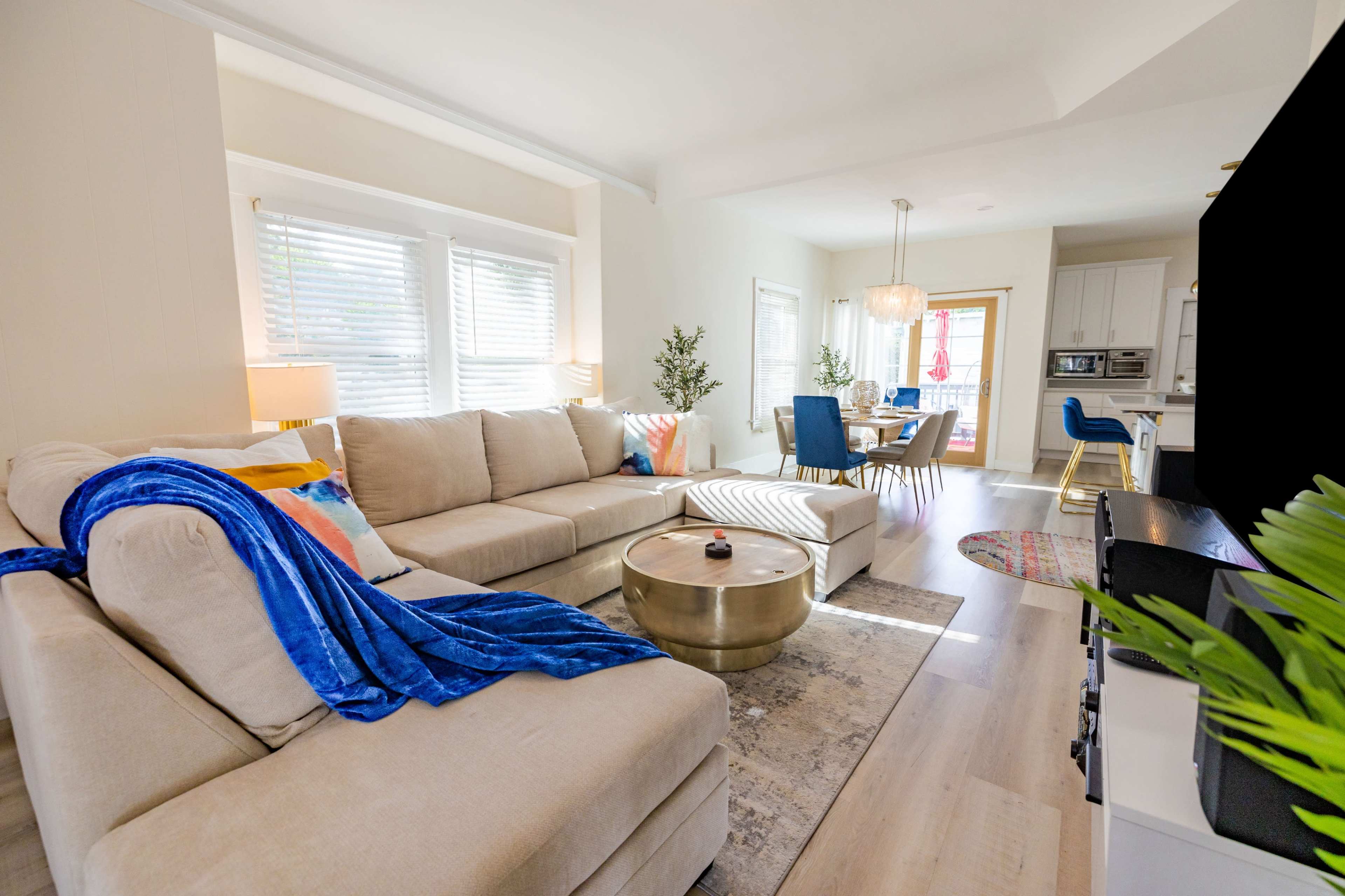 A well-furnished living room with a beige sectional sofa, a round metal coffee table, and a dining area visible in the background.