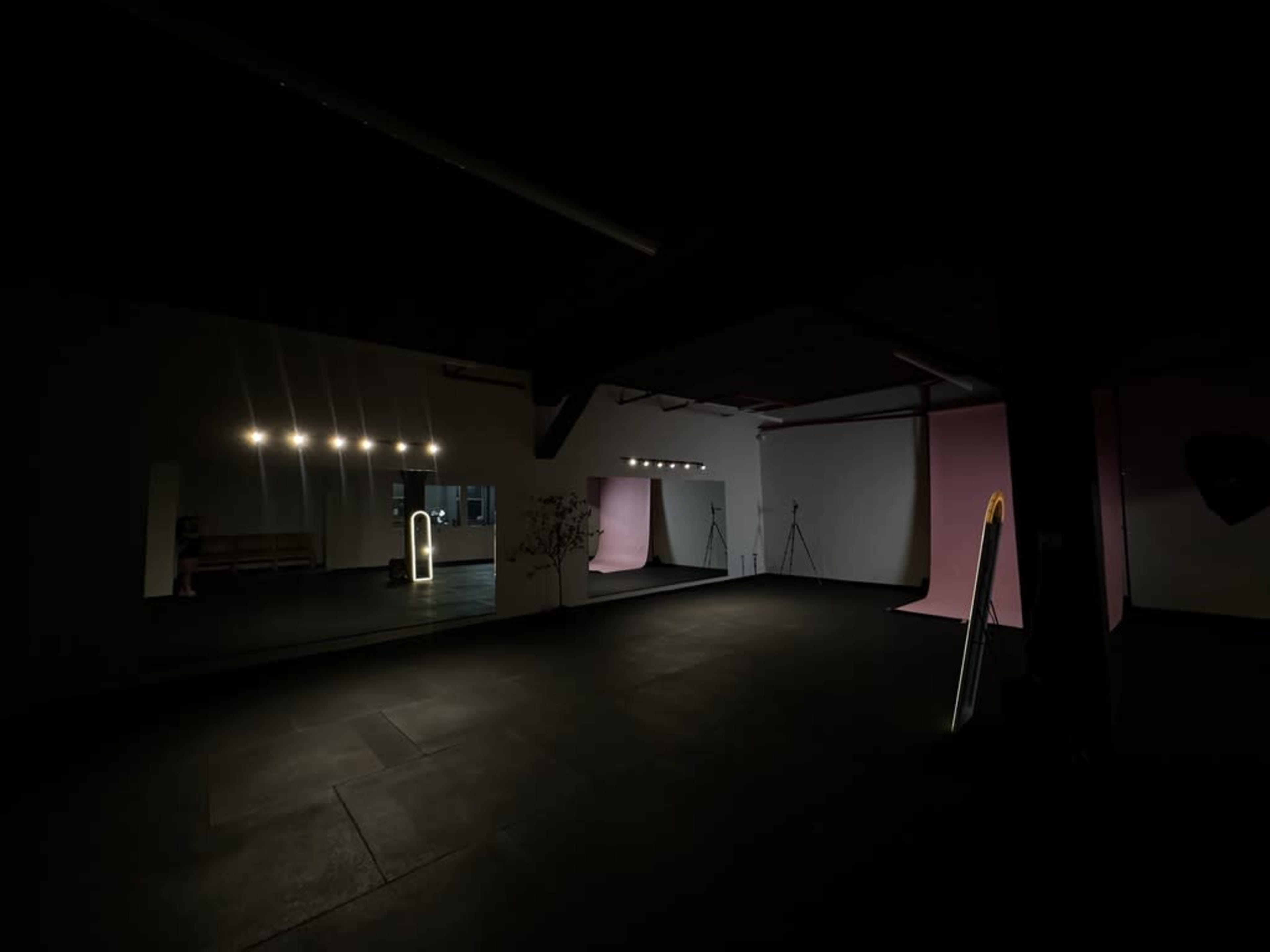 The image shows a dimly lit studio space with a large mirror, soft lighting fixtures, and a pink backdrop.