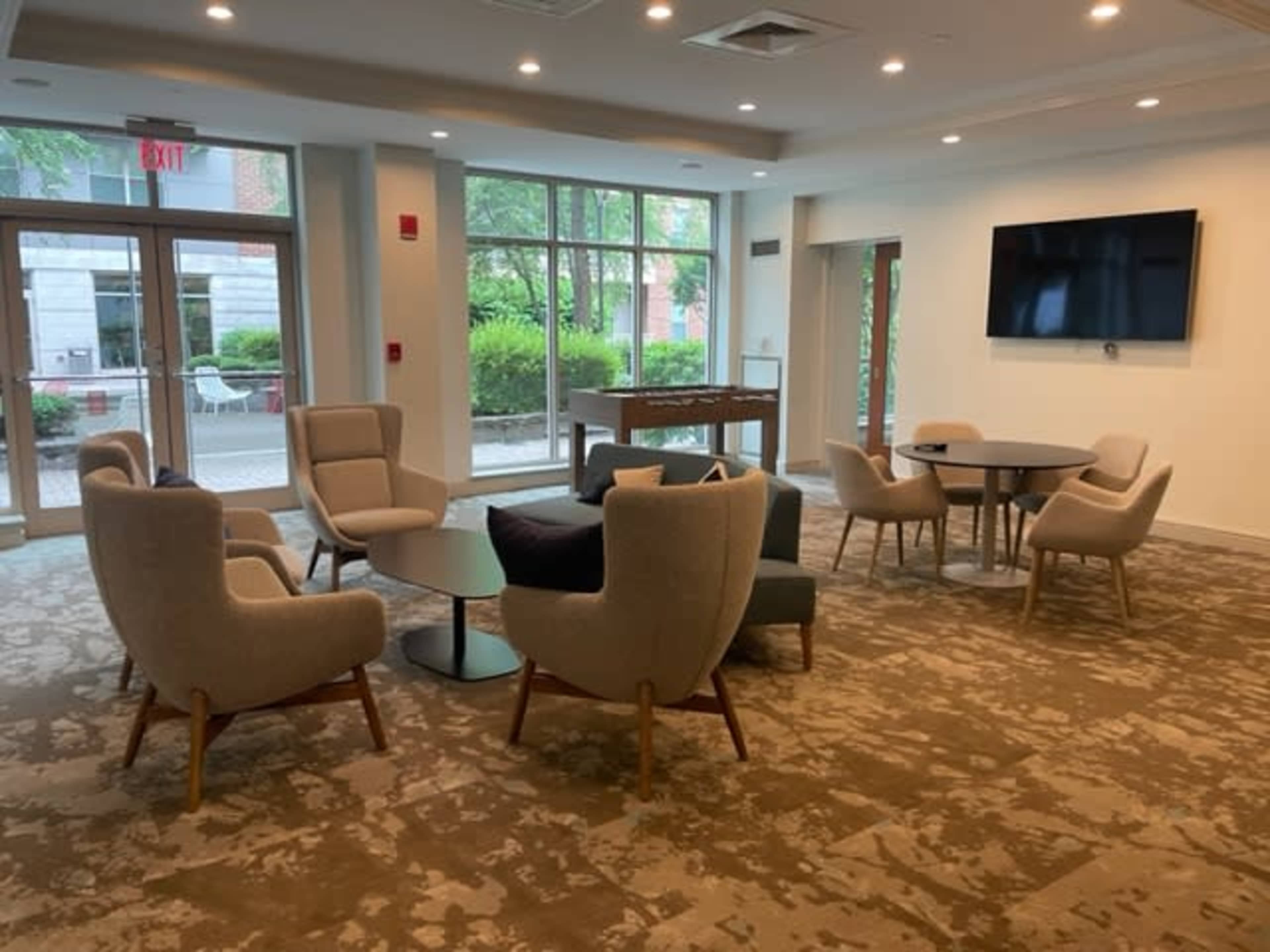 A modern lounge area features several upholstered chairs, a coffee table, and a large wall-mounted television, with windows overlooking a green outdoor space.
