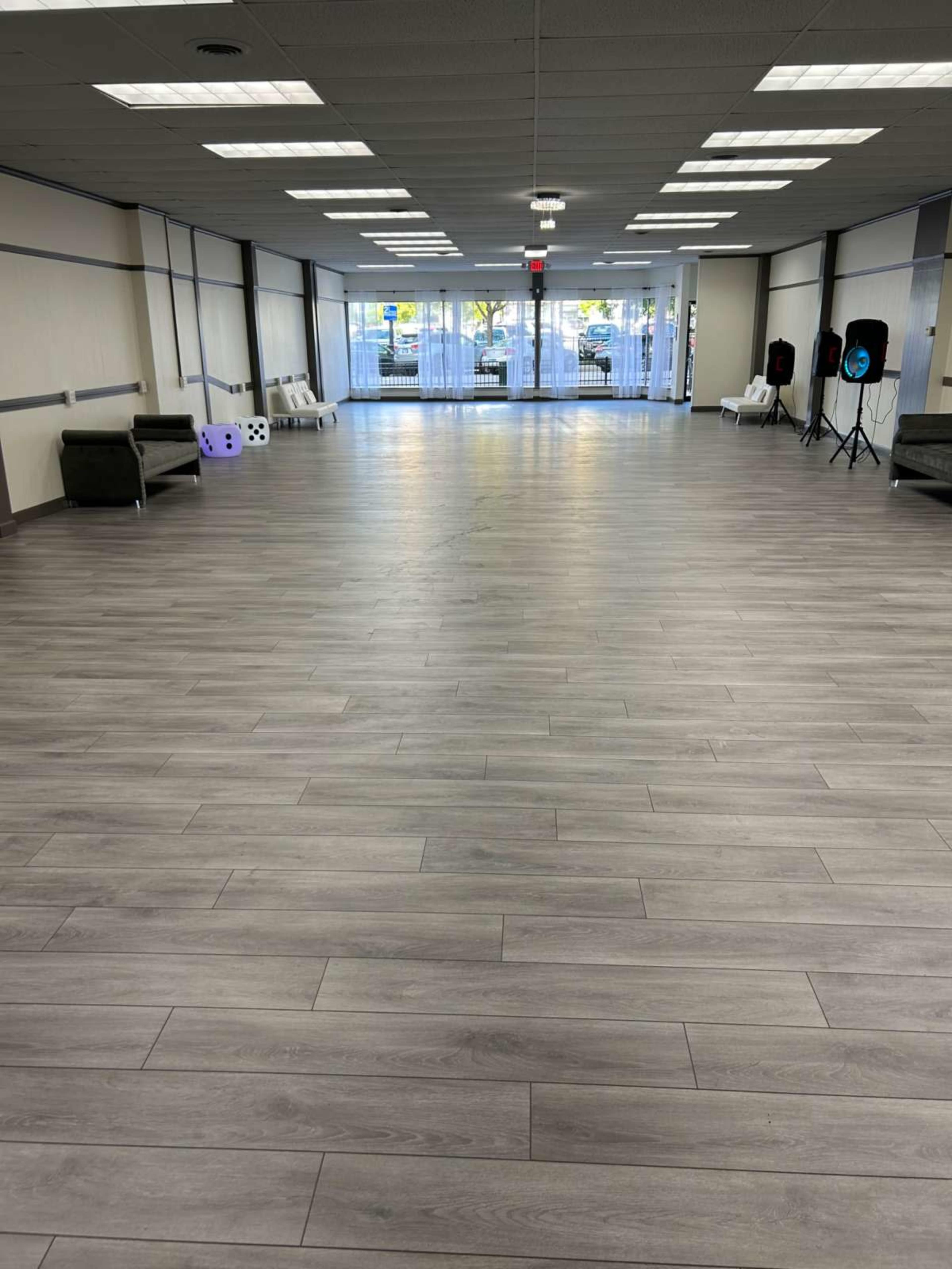 The image shows a spacious room with gray wooden flooring, empty seating areas on the sides, and large windows letting in natural light.