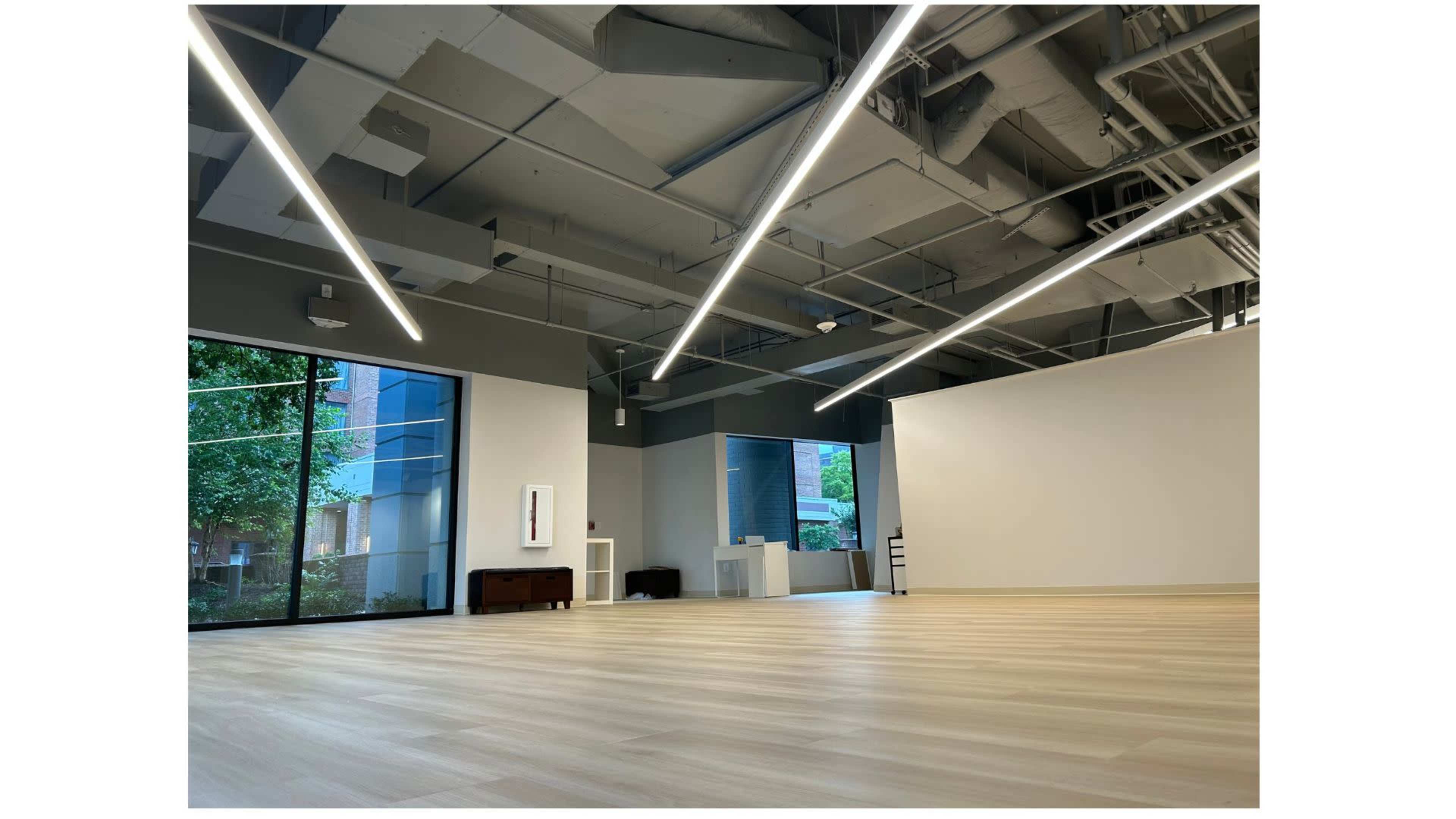 The image shows a spacious, modern interior with wooden flooring, large windows allowing natural light, and stylish ceiling lighting.