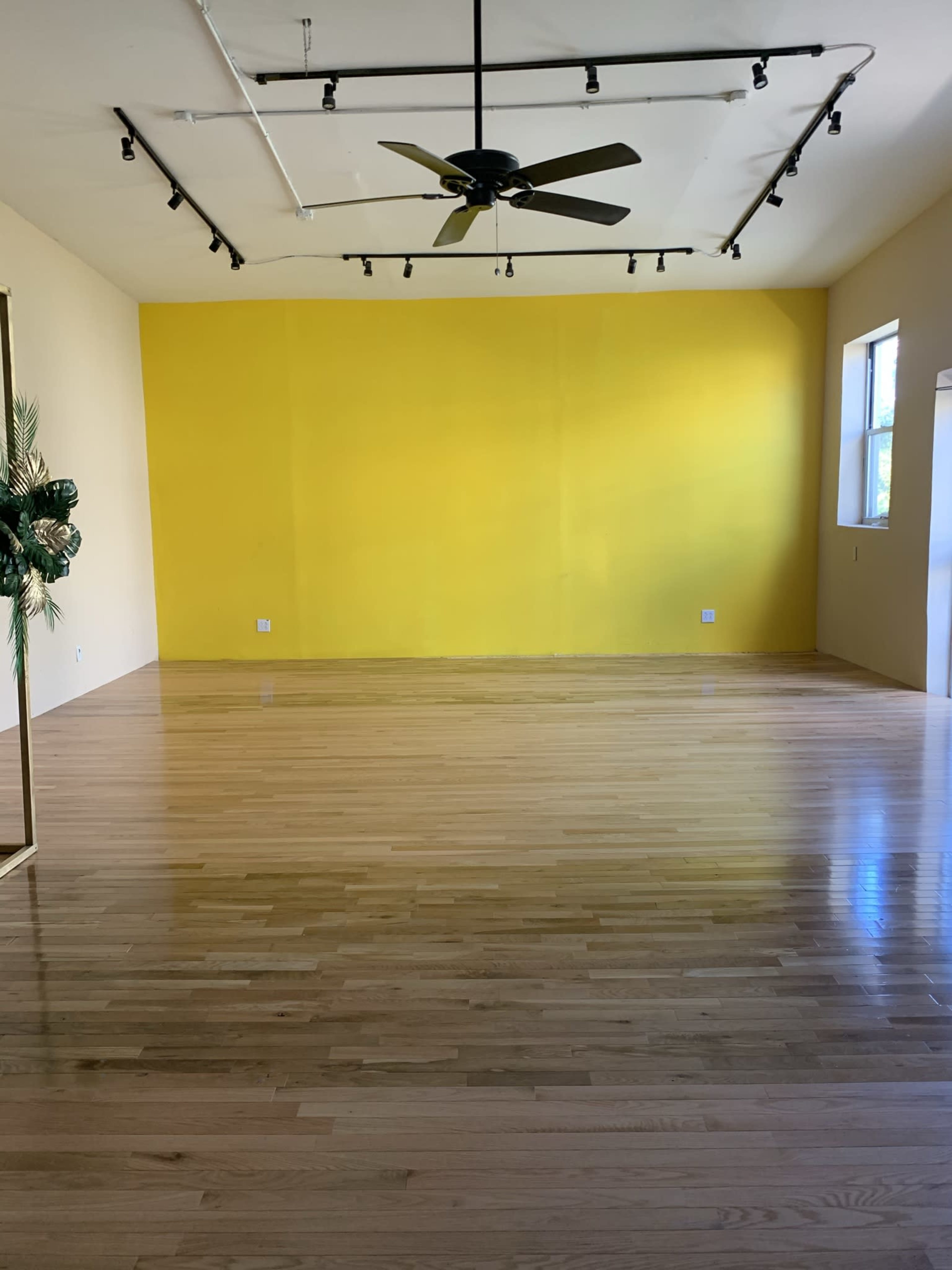 The image shows a spacious room with a yellow wall, wooden flooring, and a ceiling fan.