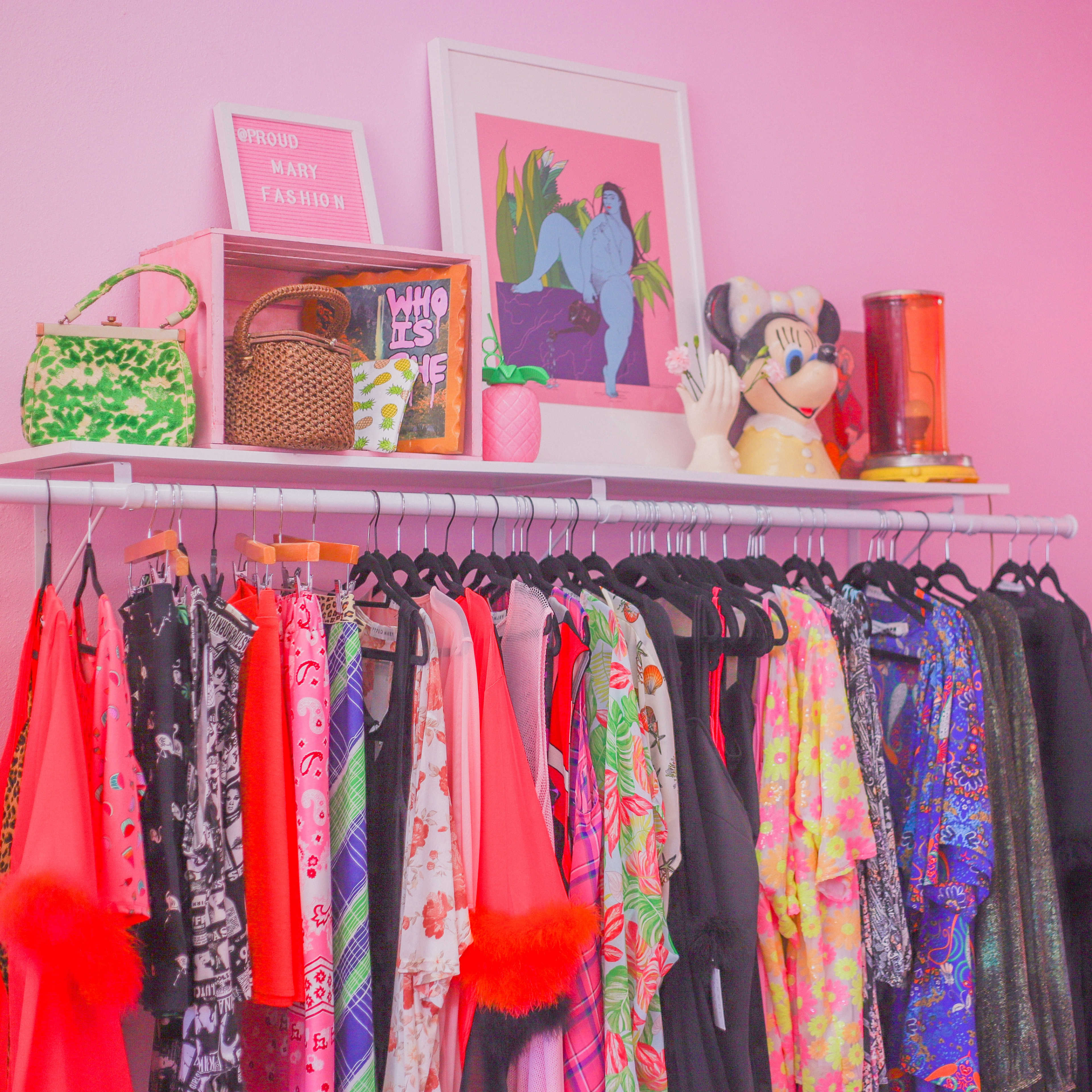 A vibrant collection of clothing hangs on a rack beneath a shelf decorated with a framed artwork, vintage bags, and a figurine against a pink wall.