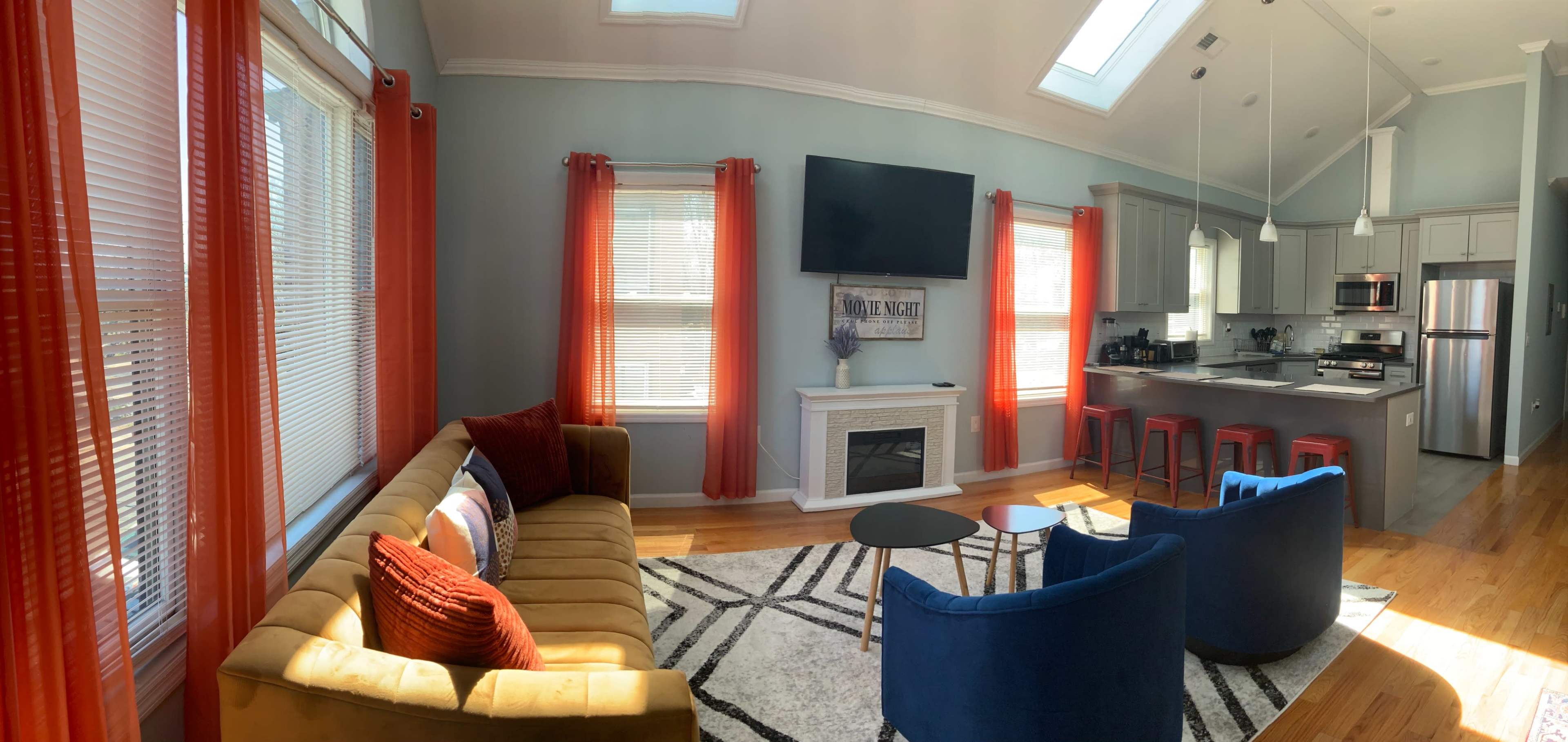 A bright living room with a yellow couch, two blue chairs, a circular black table, and a television mounted above a fireplace, all under skylights and orange curtains.