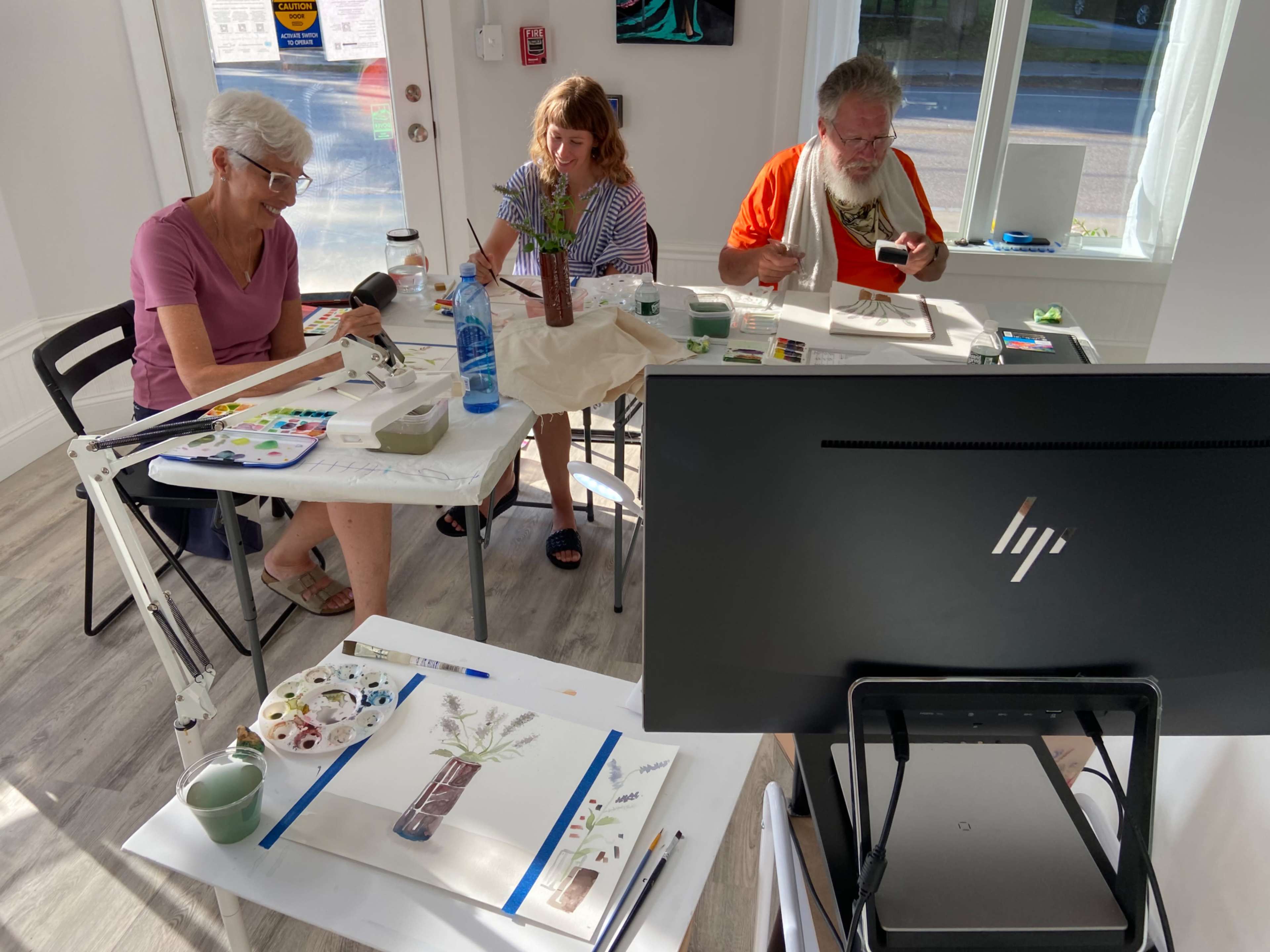 Three people are seated at a table engaged in a painting class, surrounded by art supplies and natural light coming through a large window.