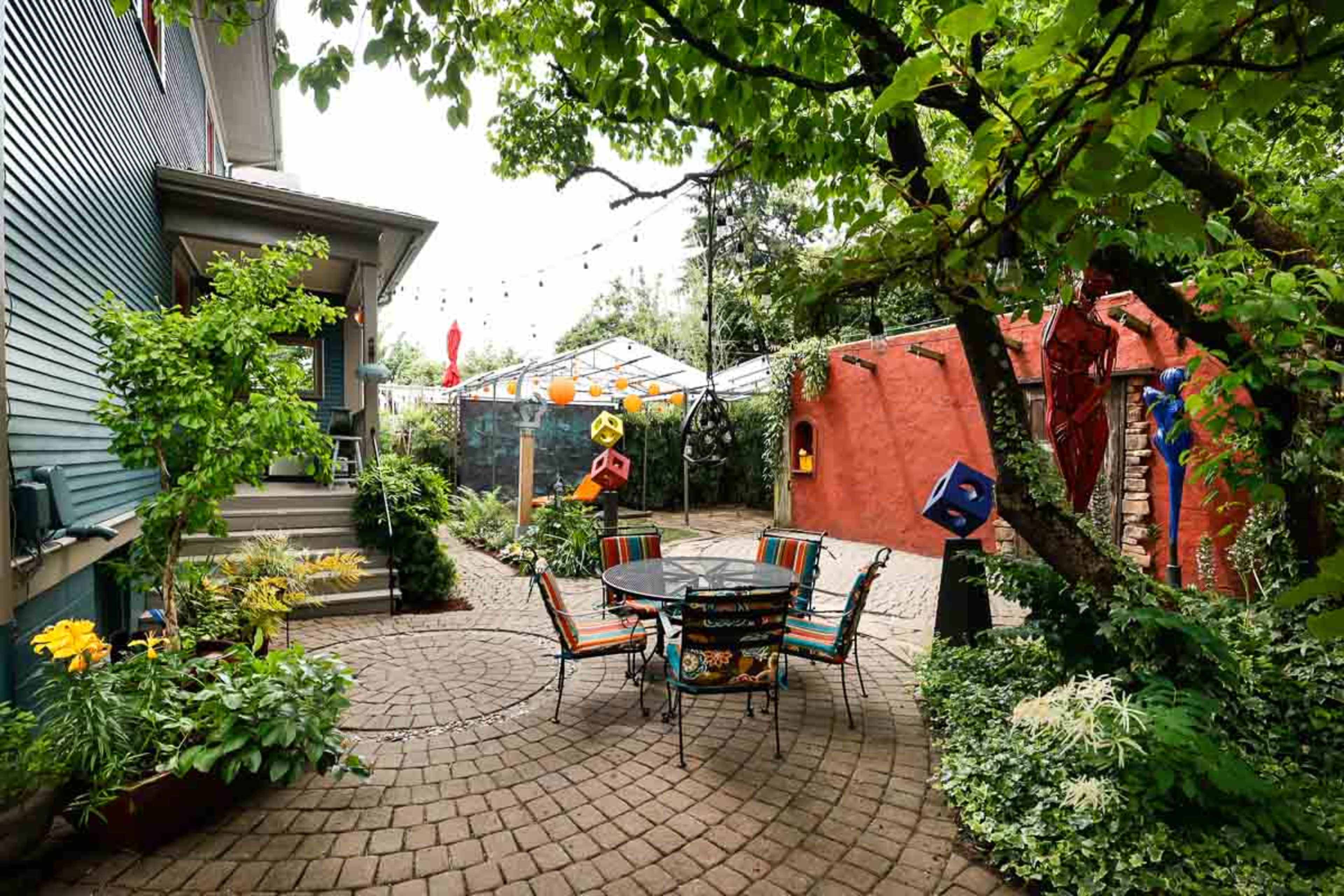A circular stone patio features a black metal table and chairs surrounded by lush greenery and colorful decorations.