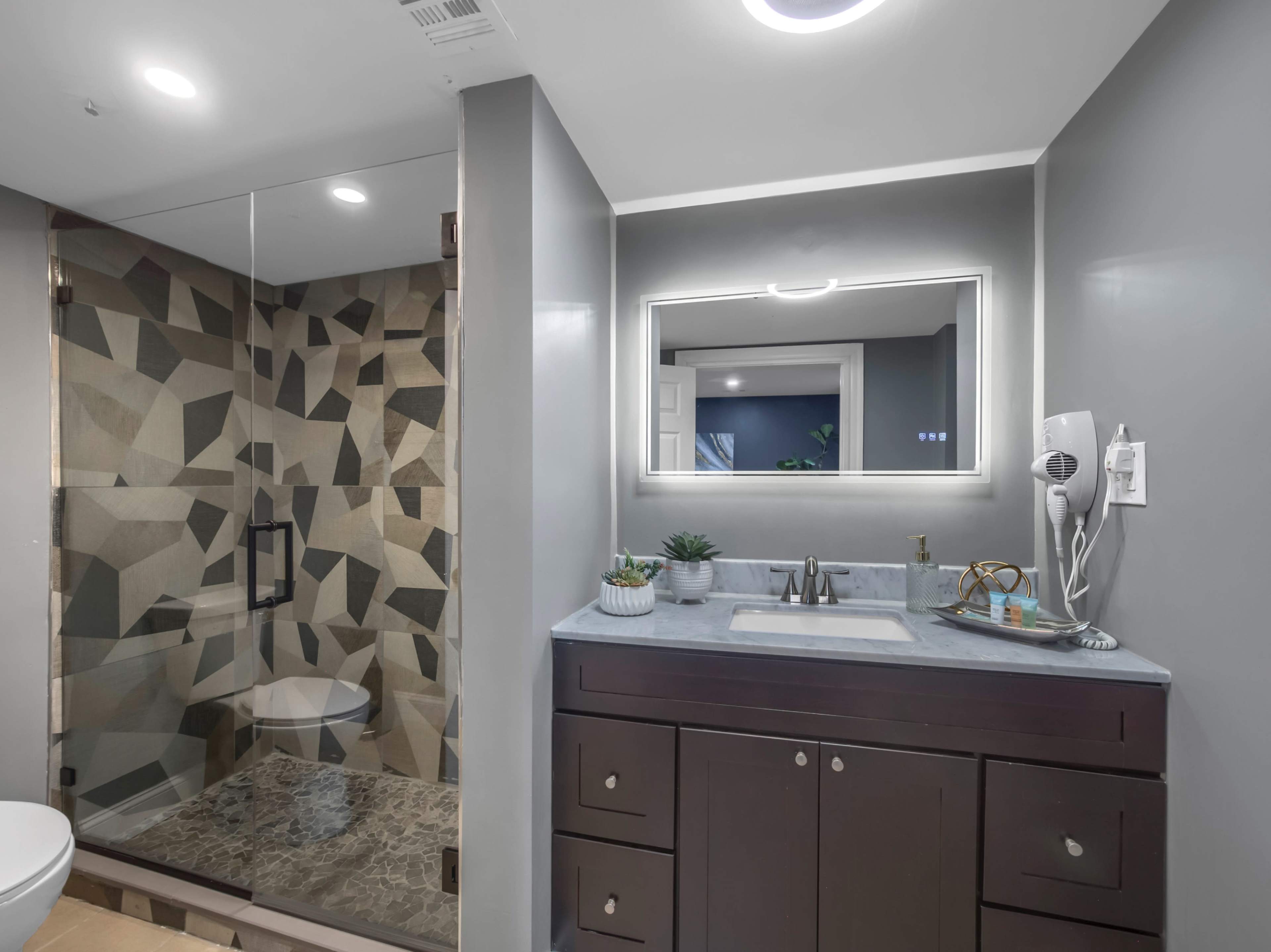 The bathroom features a glass-enclosed shower with geometric tile patterns, a modern vanity with a sink, and a backlit mirror.