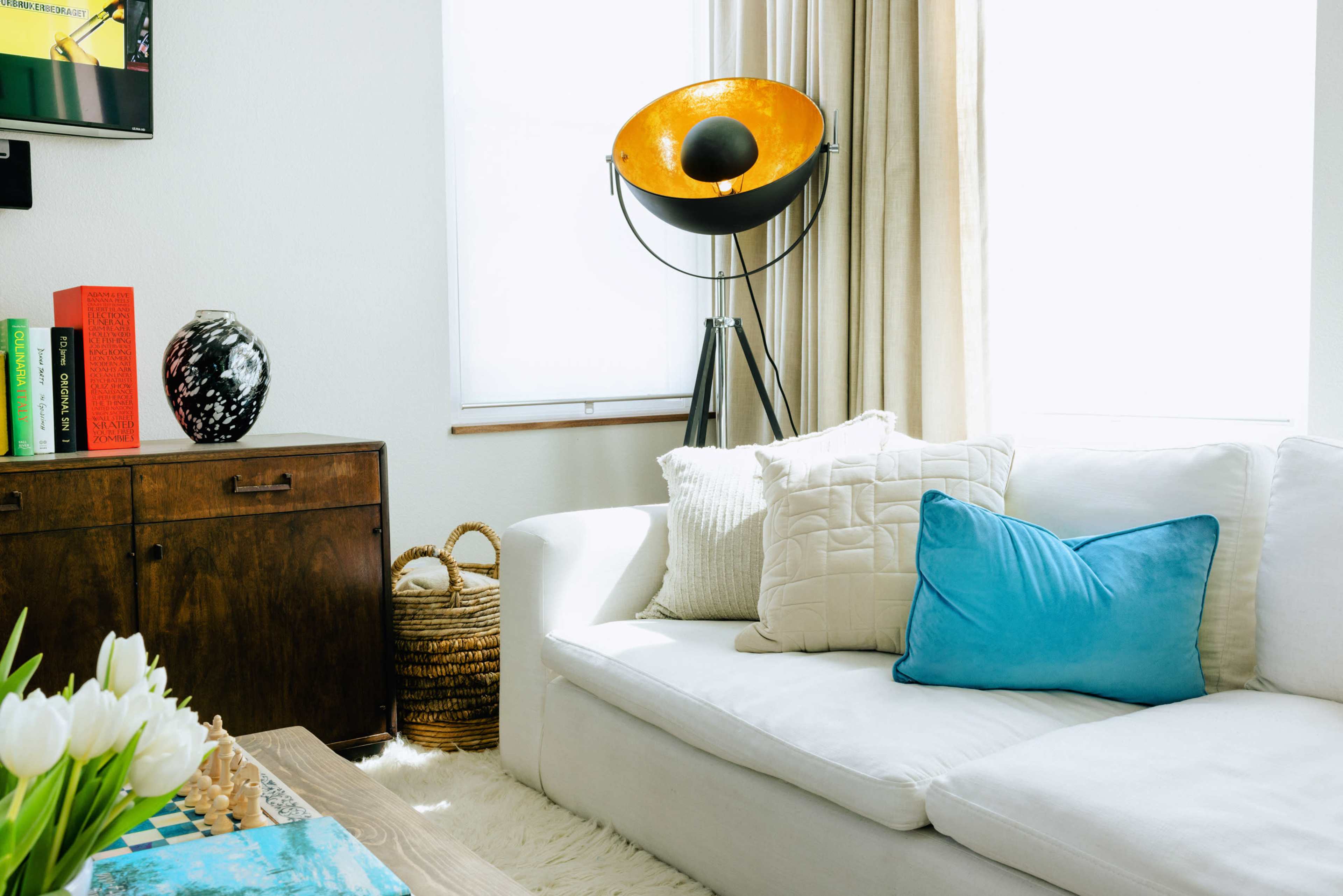 A bright living room features a white sofa with assorted cushions, a wooden cabinet, and a floor lamp with a large orange shade.