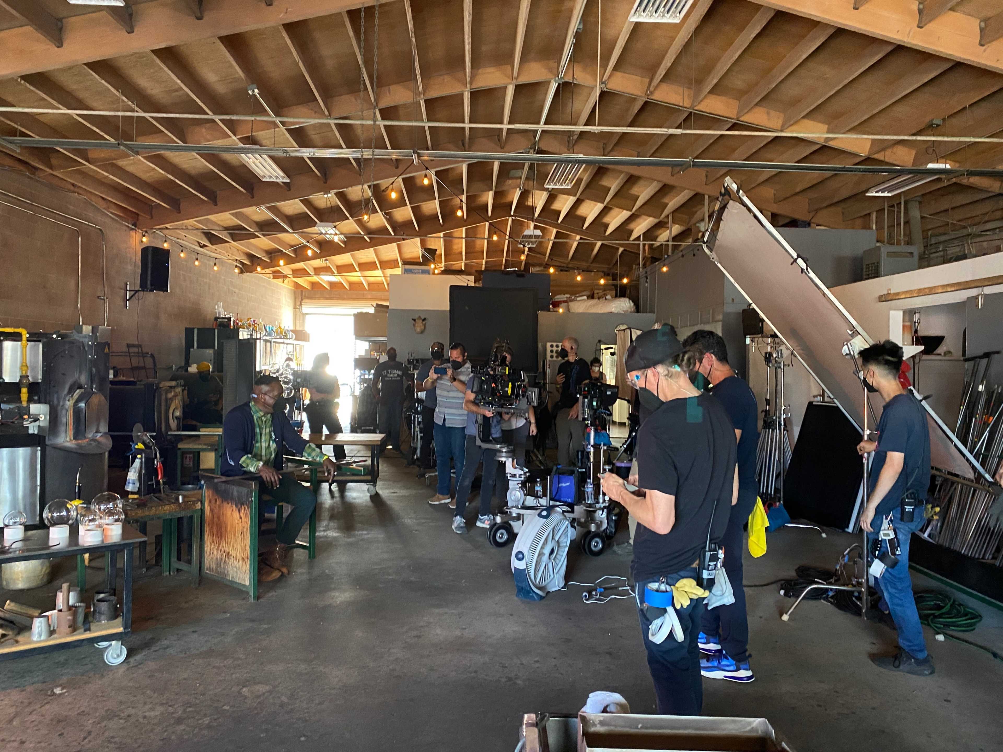 A film crew sets up equipment in a large industrial workshop space.
