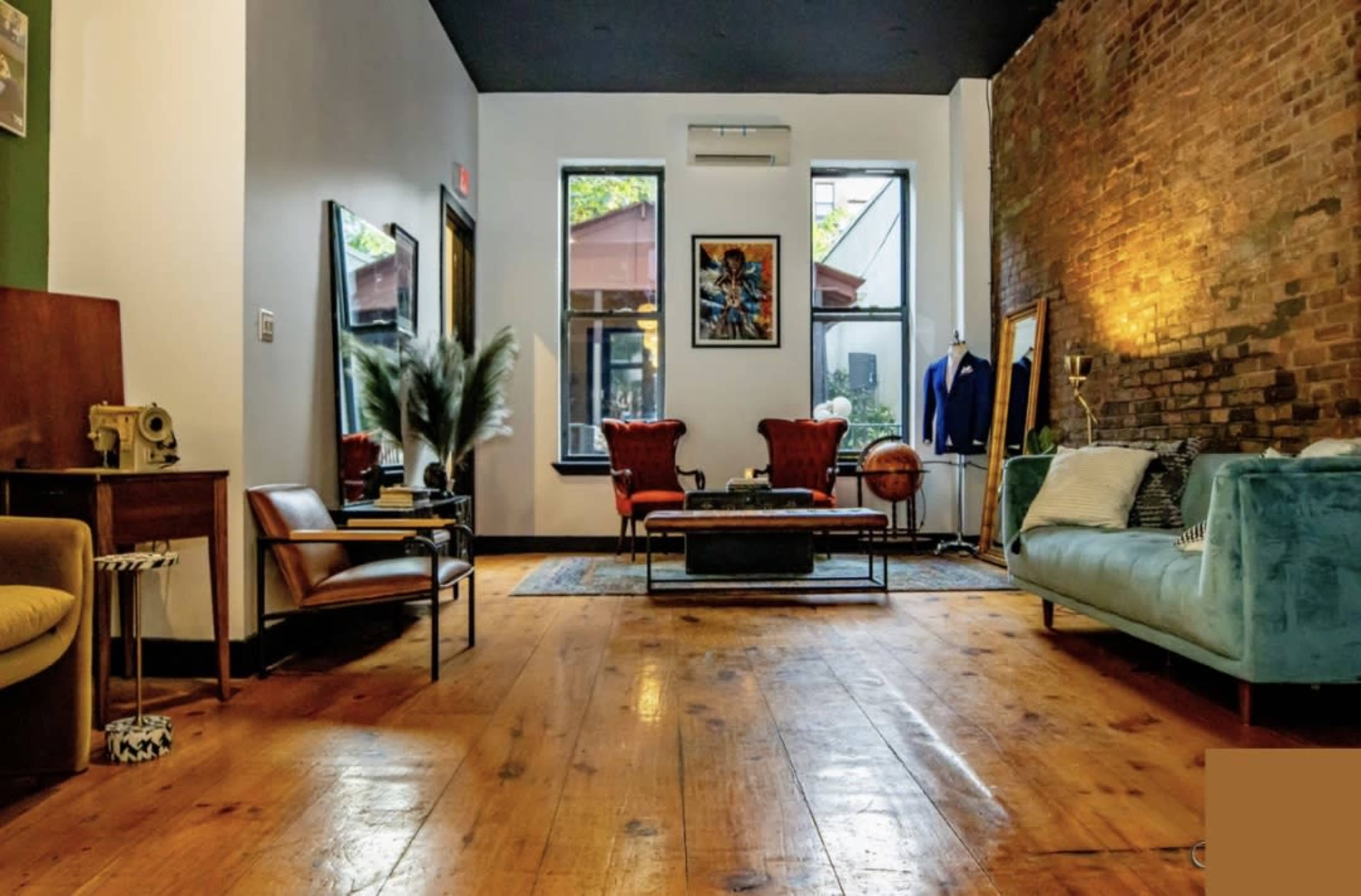 The image shows a well-furnished room with exposed brick walls, wooden flooring, and a mixture of seating options, including a teal couch and red armchairs.