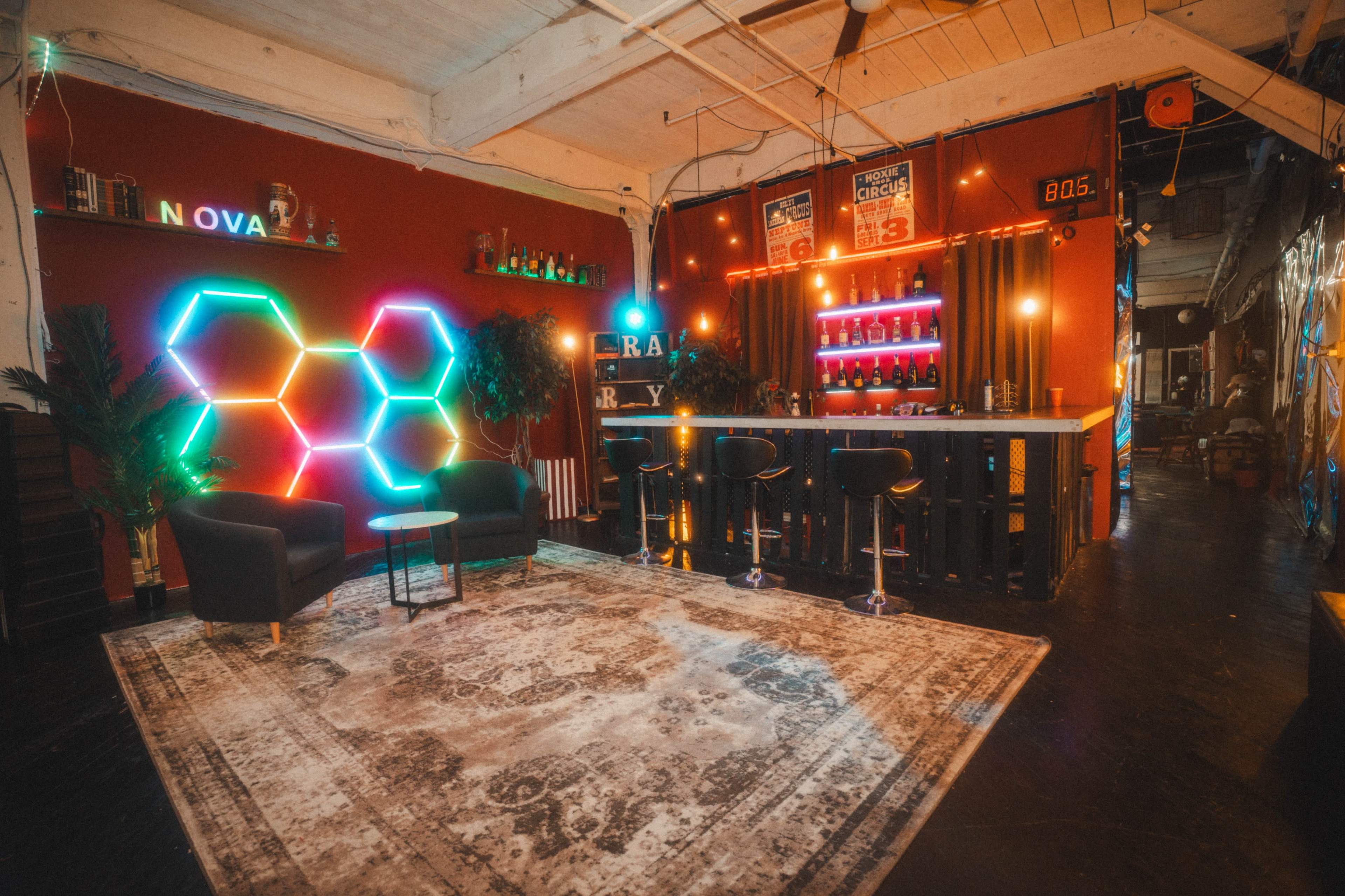 The image shows a modern lounge area featuring a bar with colorful neon lighting, two black chairs, and a patterned rug on a dark floor.
