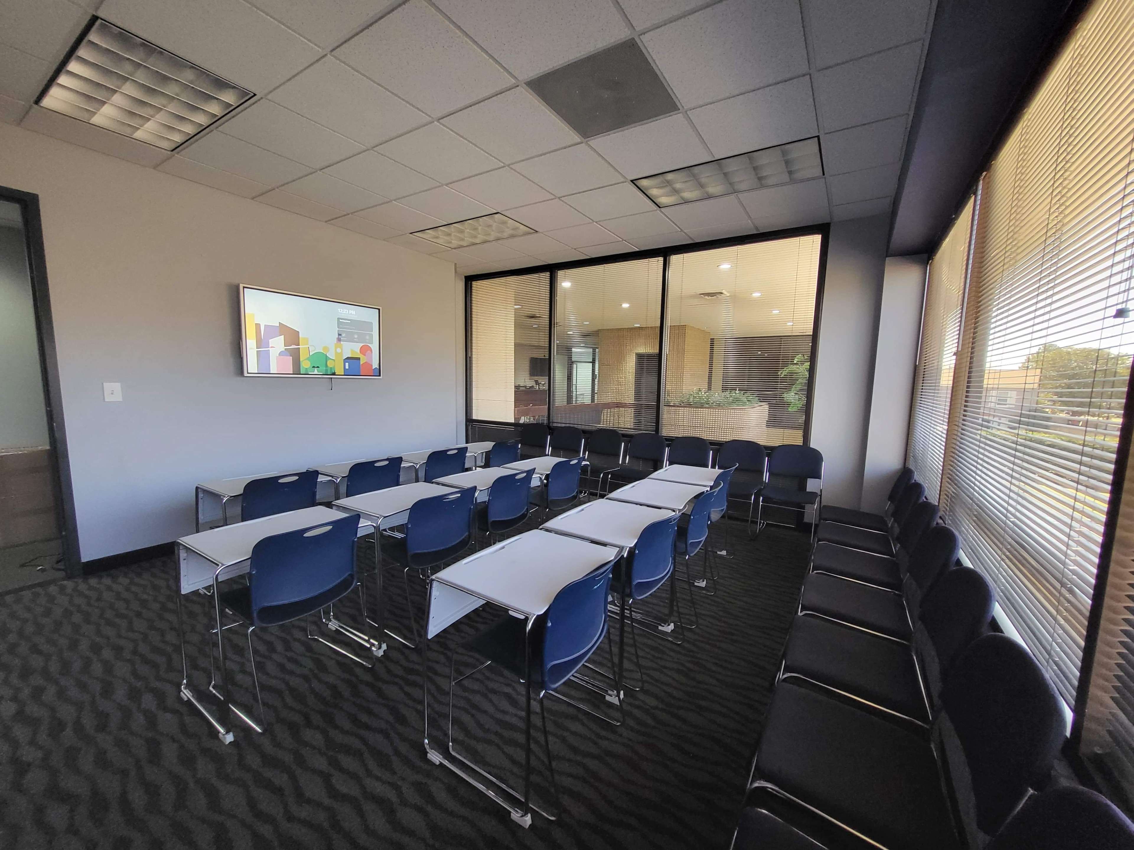 A conference room with rows of rectangular tables and blue chairs facing a wall-mounted screen, with large windows overlooking a building's interior.