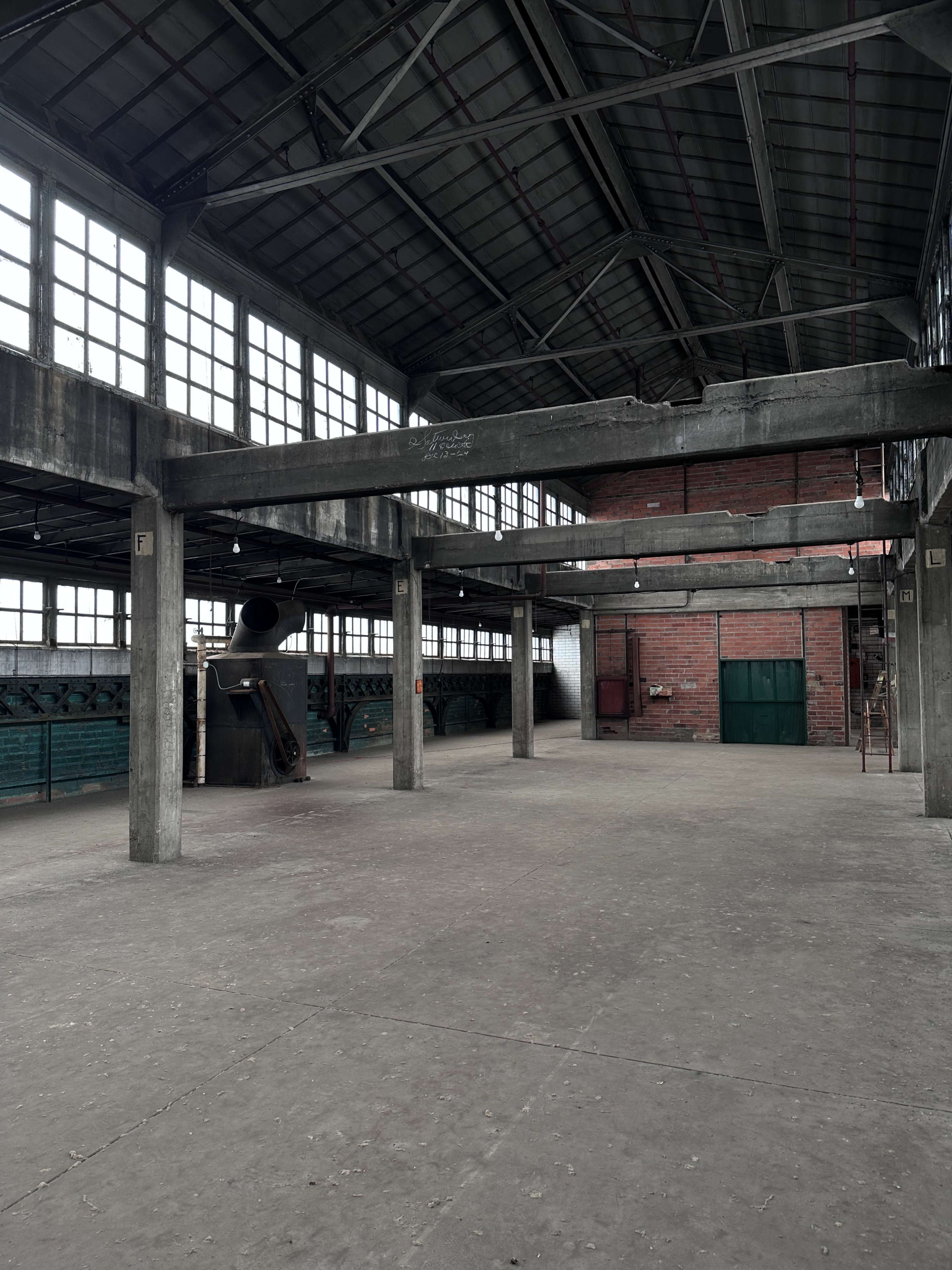 The image shows a large, empty industrial space with high ceilings, large windows, and concrete support beams.