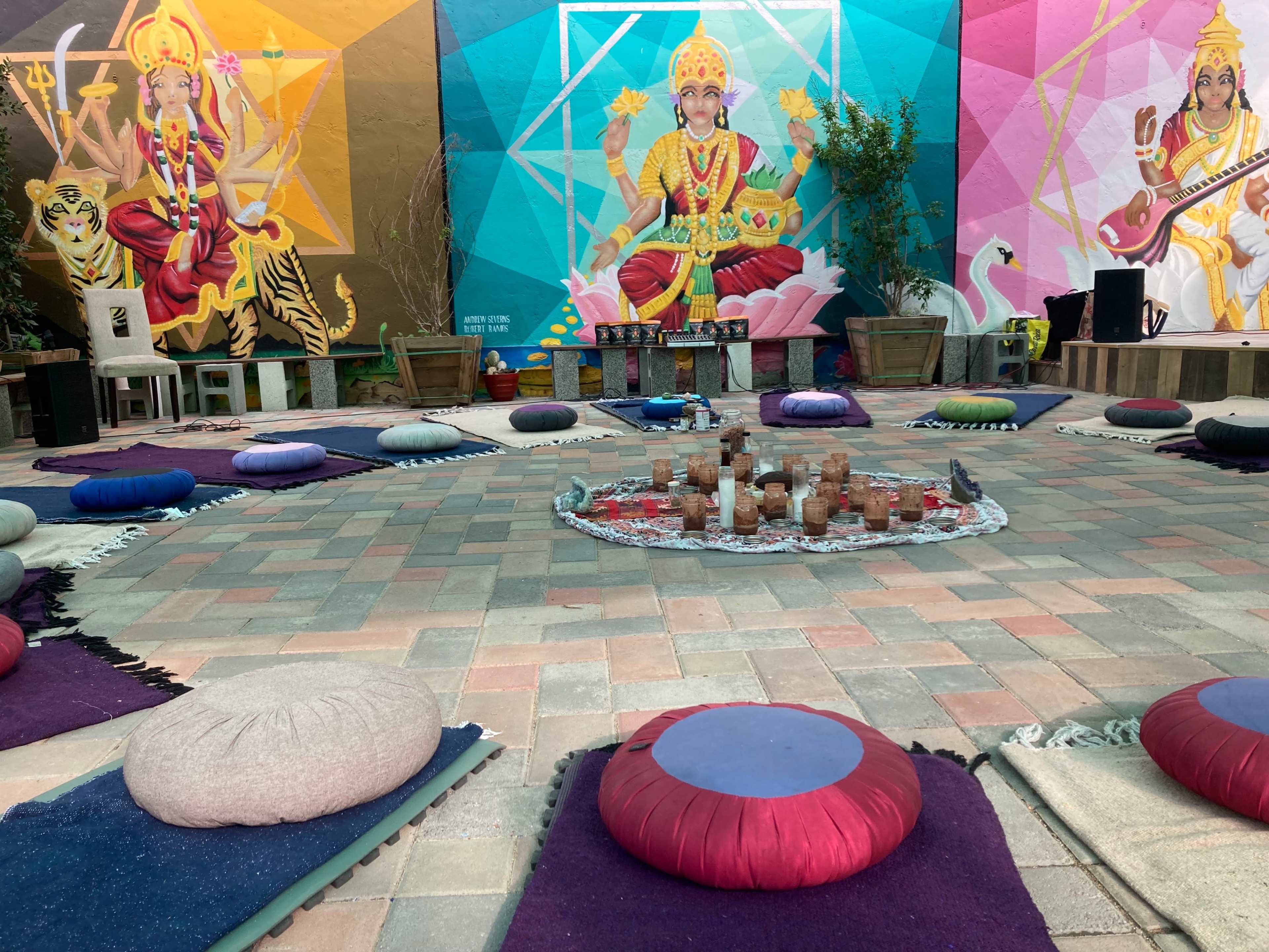A colorful outdoor space arranged with circular seating cushions surrounding a central arrangement of candles and decorative items, set against vibrant murals of deities.