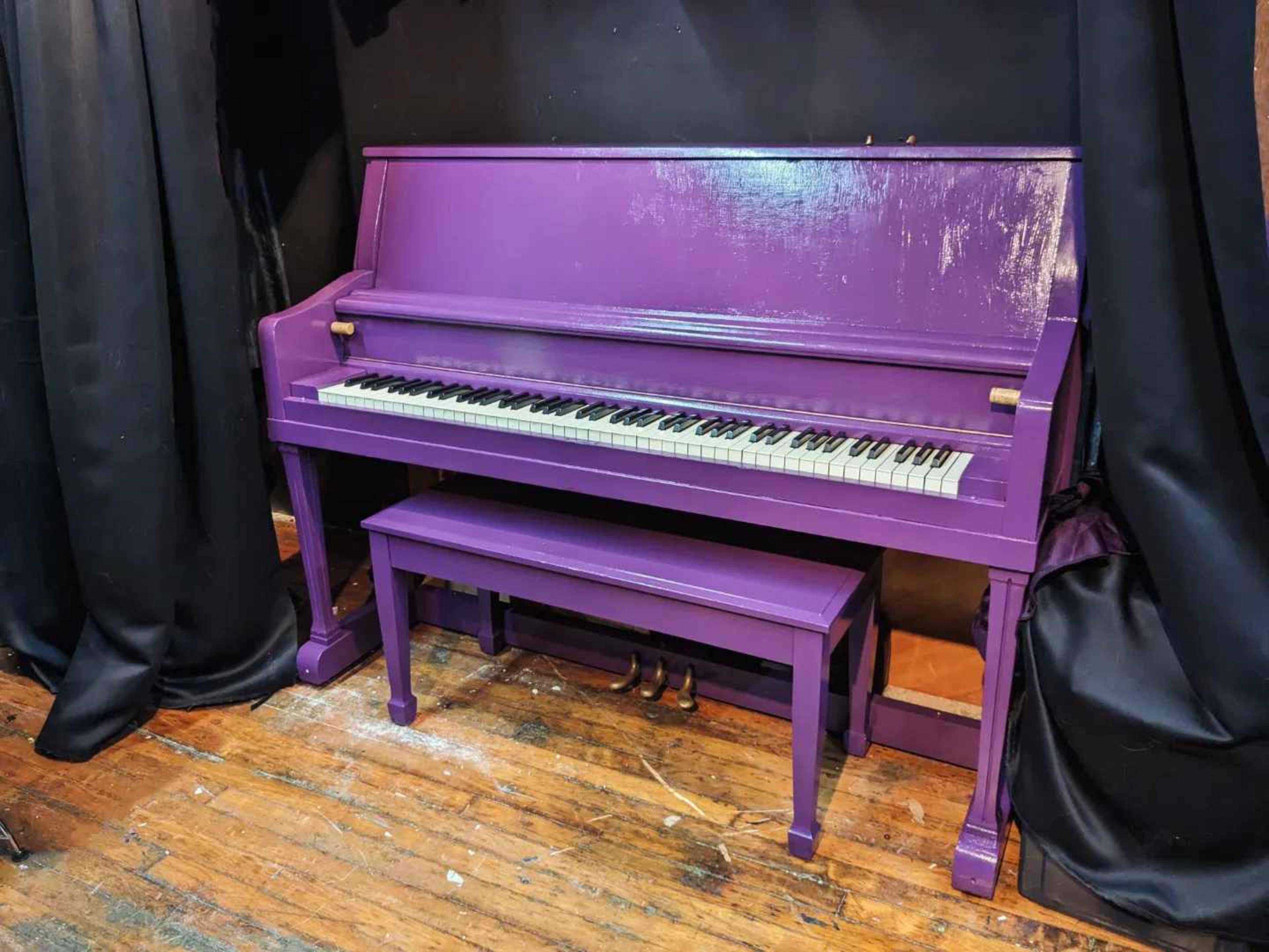 A purple piano and matching bench are positioned against a black backdrop in a dimly lit room.