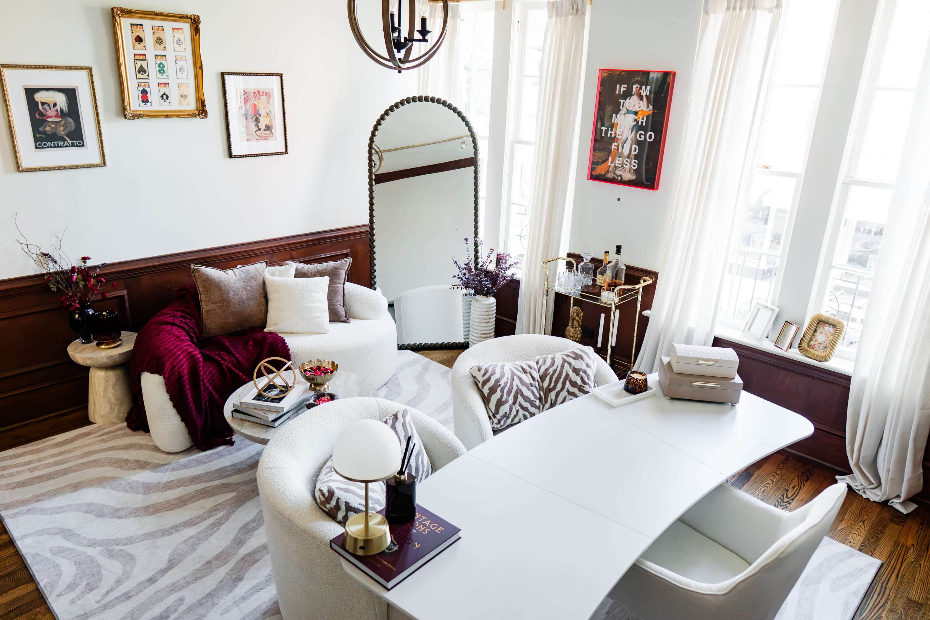 The image shows a modern living room featuring plush seating, a sleek white desk, decorative art on the walls, and large windows with natural light.