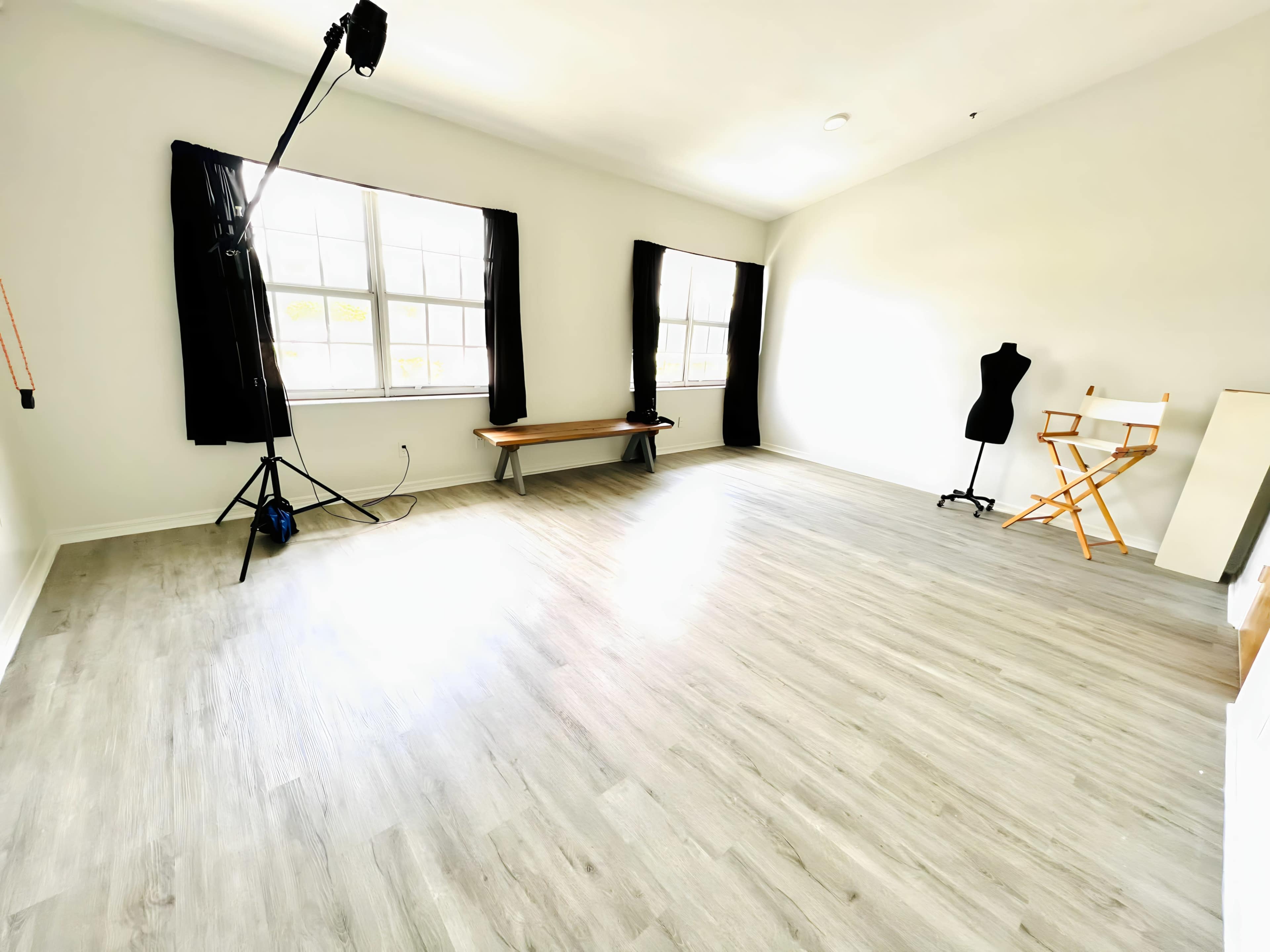 A well-lit room with light-colored flooring, two large windows with black curtains, a wooden bench, a chair, and a dress form.