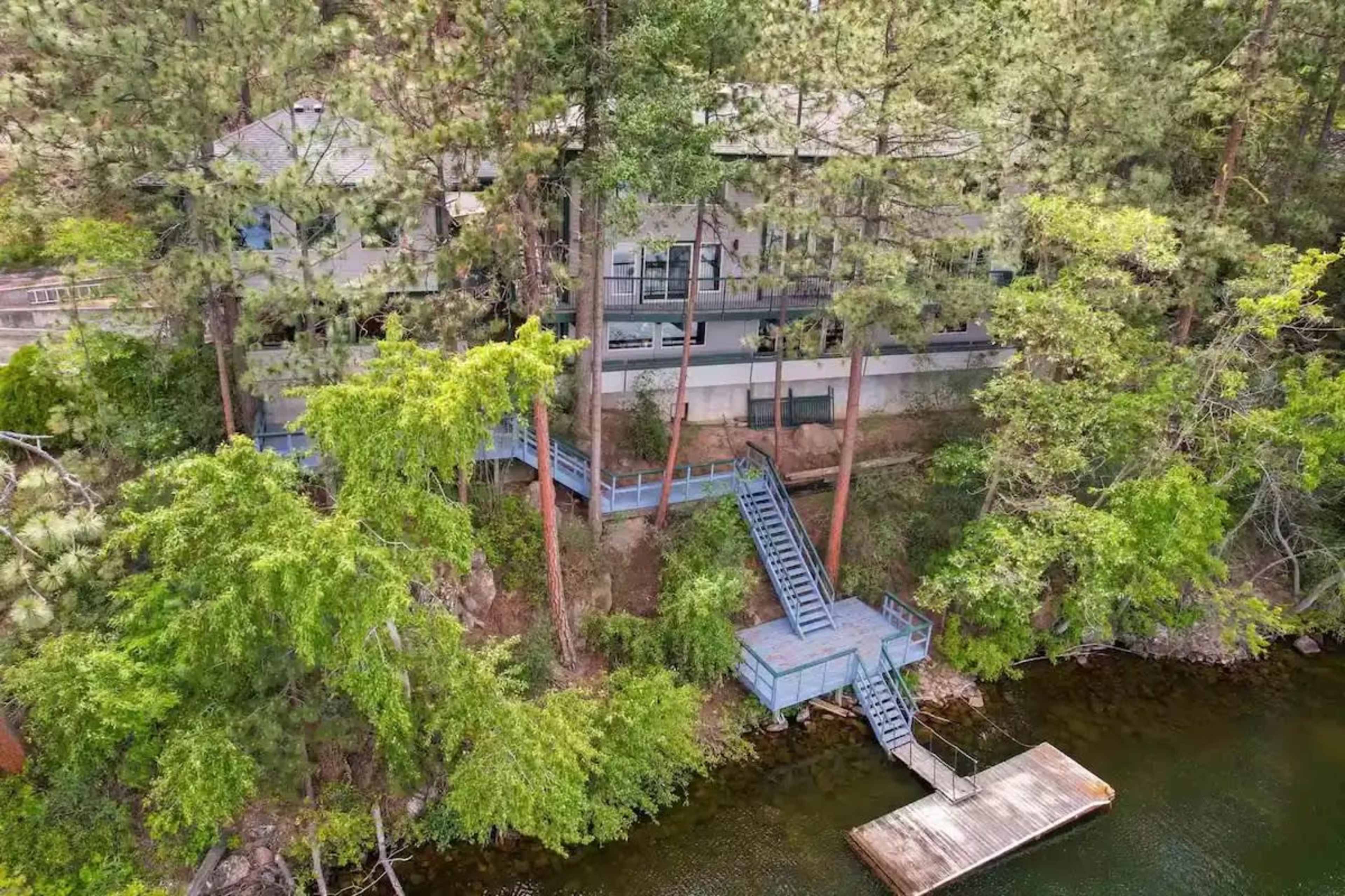 A three-story house is situated on a hillside next to a lake, surrounded by tall trees and featuring a staircase leading down to a dock.