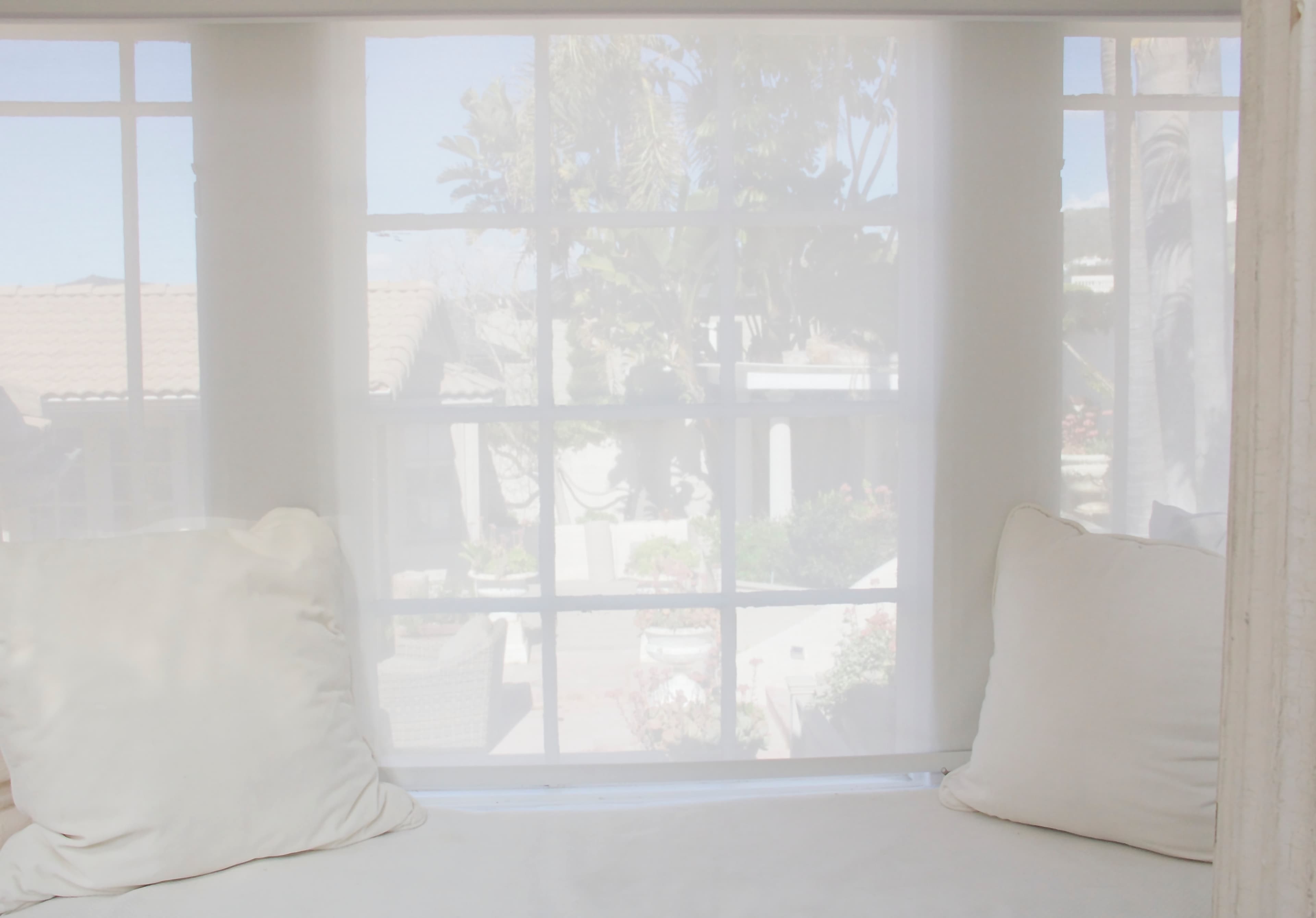 A sheer curtain partially obscures a view of an outdoor garden through a window, with two light-colored pillows resting on a bench nearby.