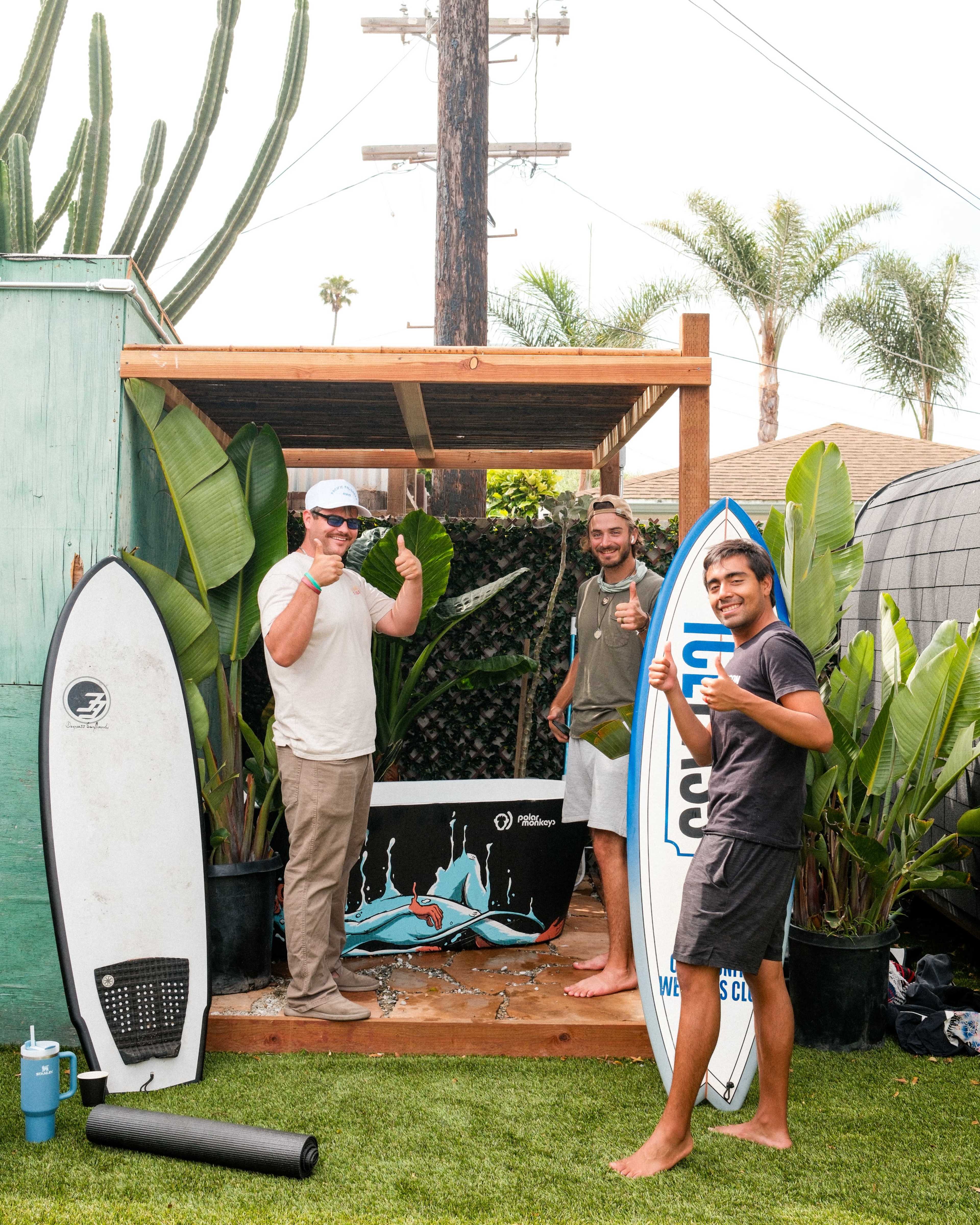 Three men pose with surfboards in a backyard surrounded by tropical plants and a wooden deck.