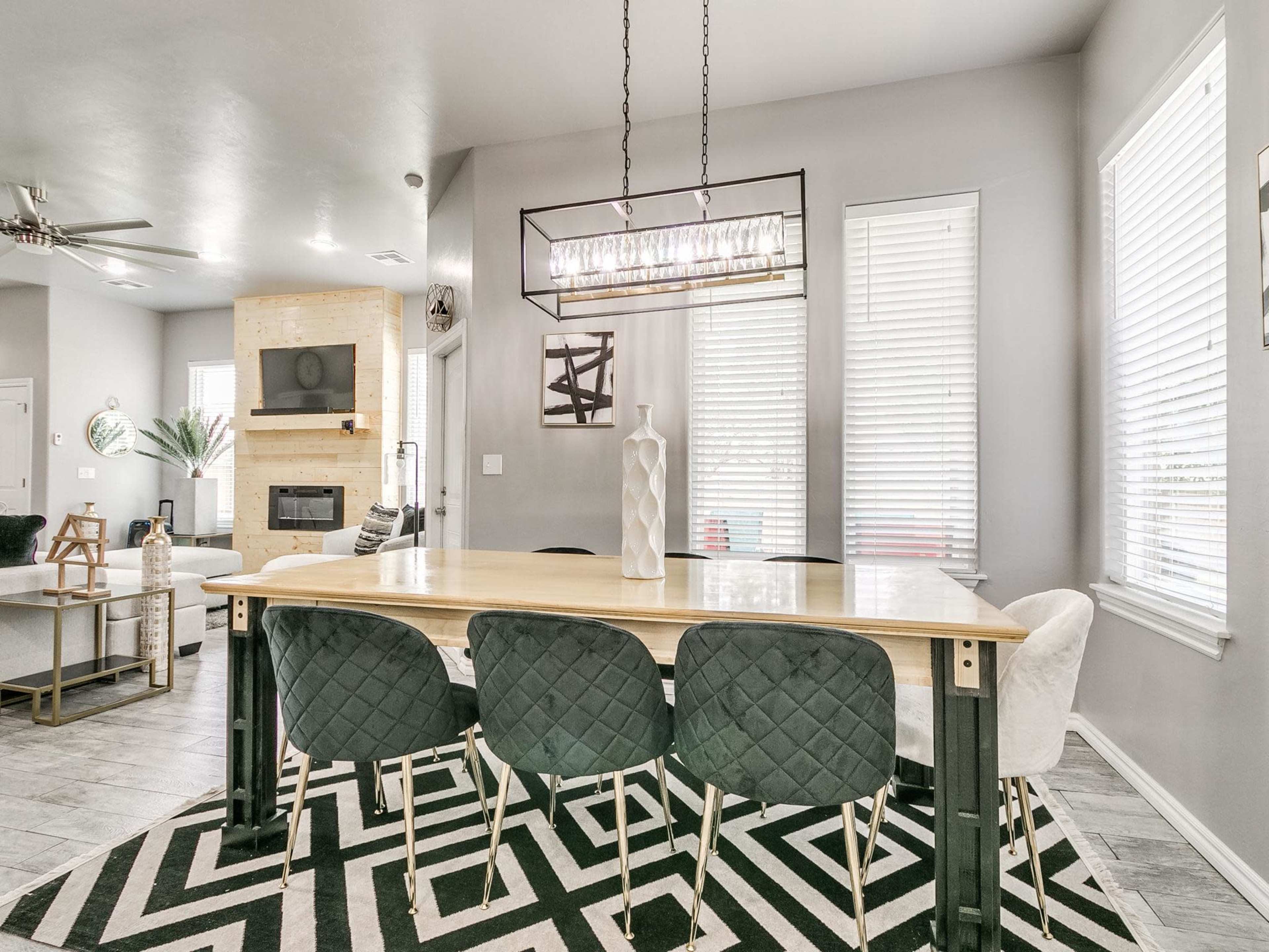 A modern dining area with a large wooden table surrounded by green upholstered chairs, featuring a geometric black and white rug and large windows providing natural light.