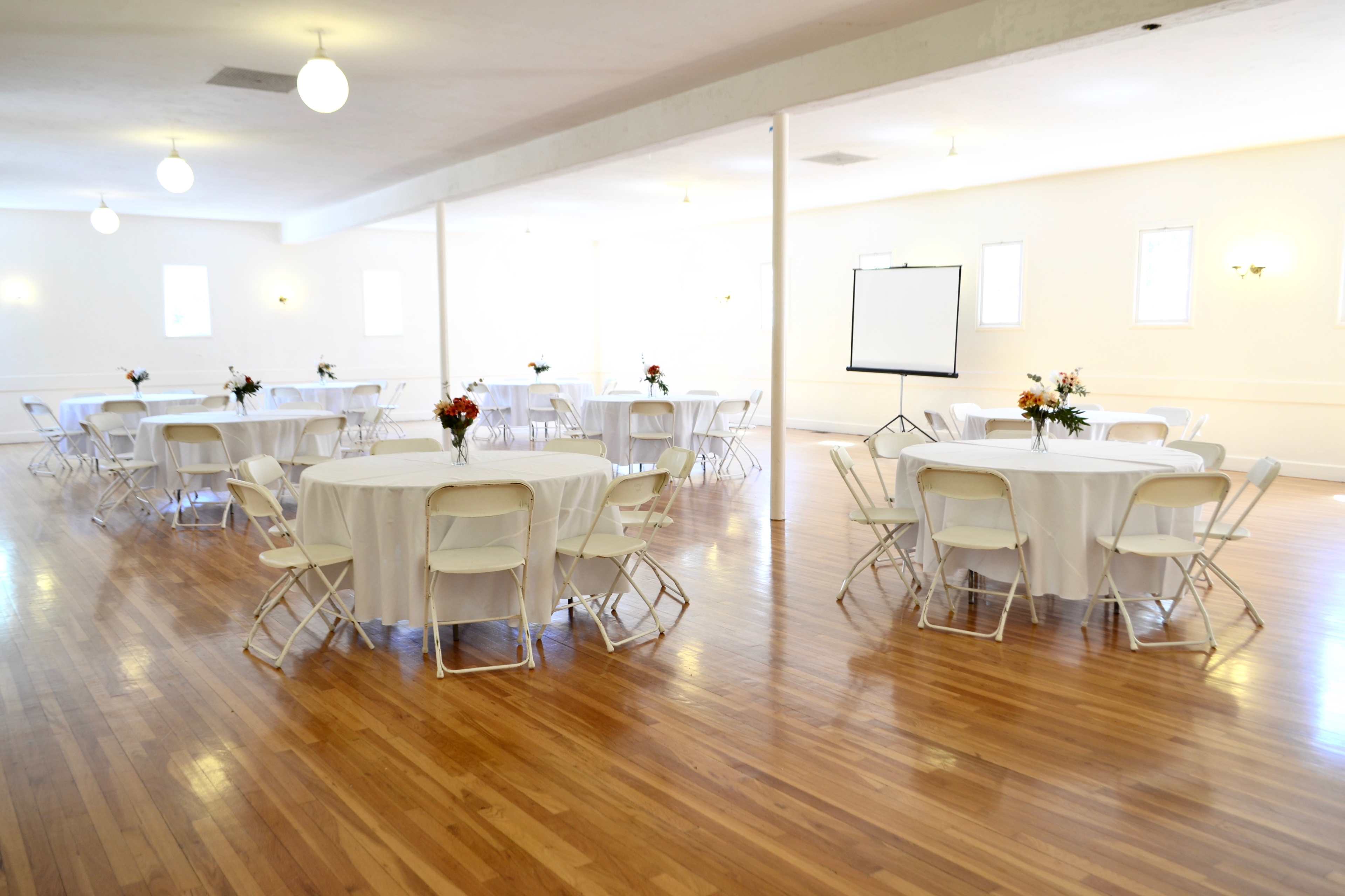 A spacious reception hall with several round tables covered in white linens, surrounded by white folding chairs, and featuring a blank easel on one side.