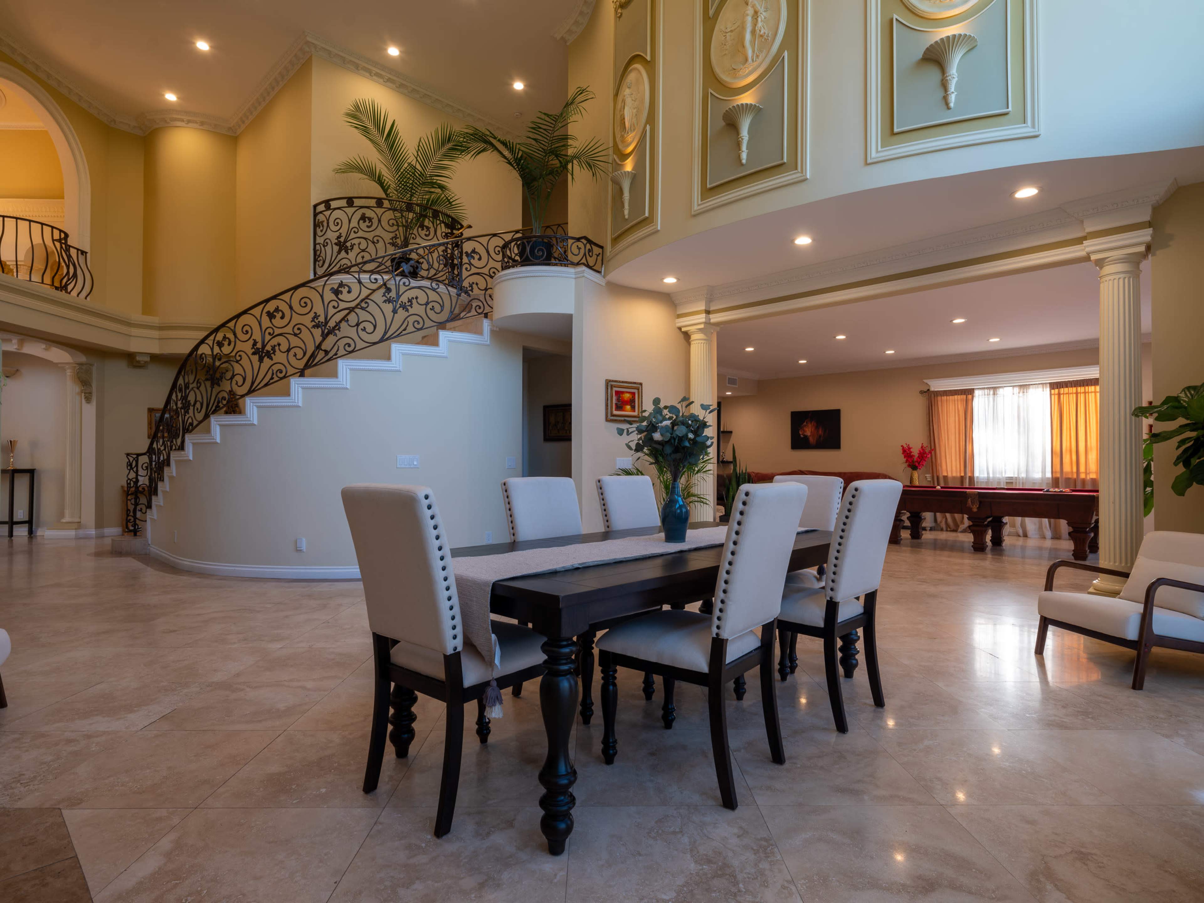A spacious dining area features a large table surrounded by chairs, with a decorative staircase and plants in the background.