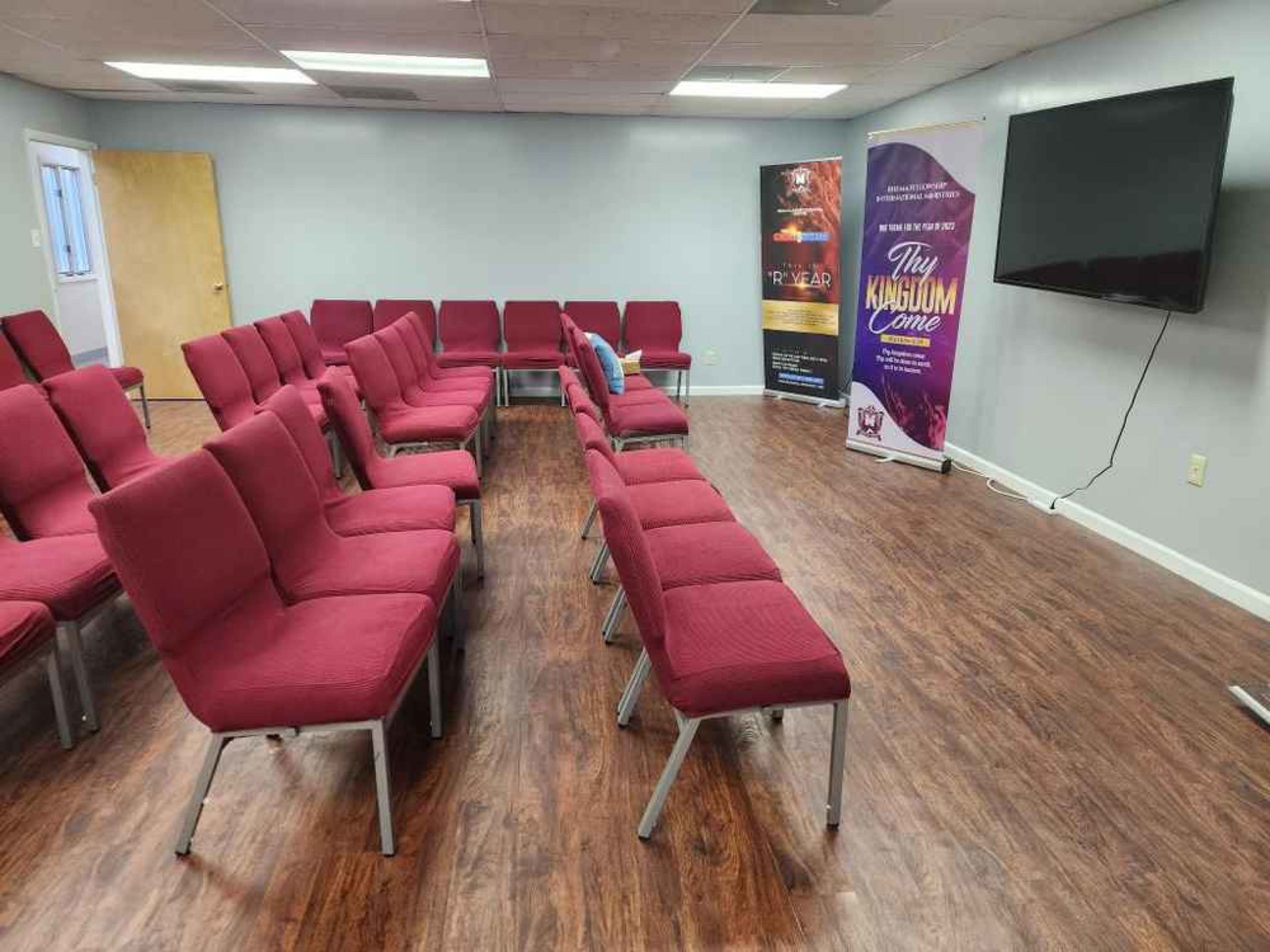 A row of red chairs is arranged in a room with a TV screen and promotional banners on the walls.