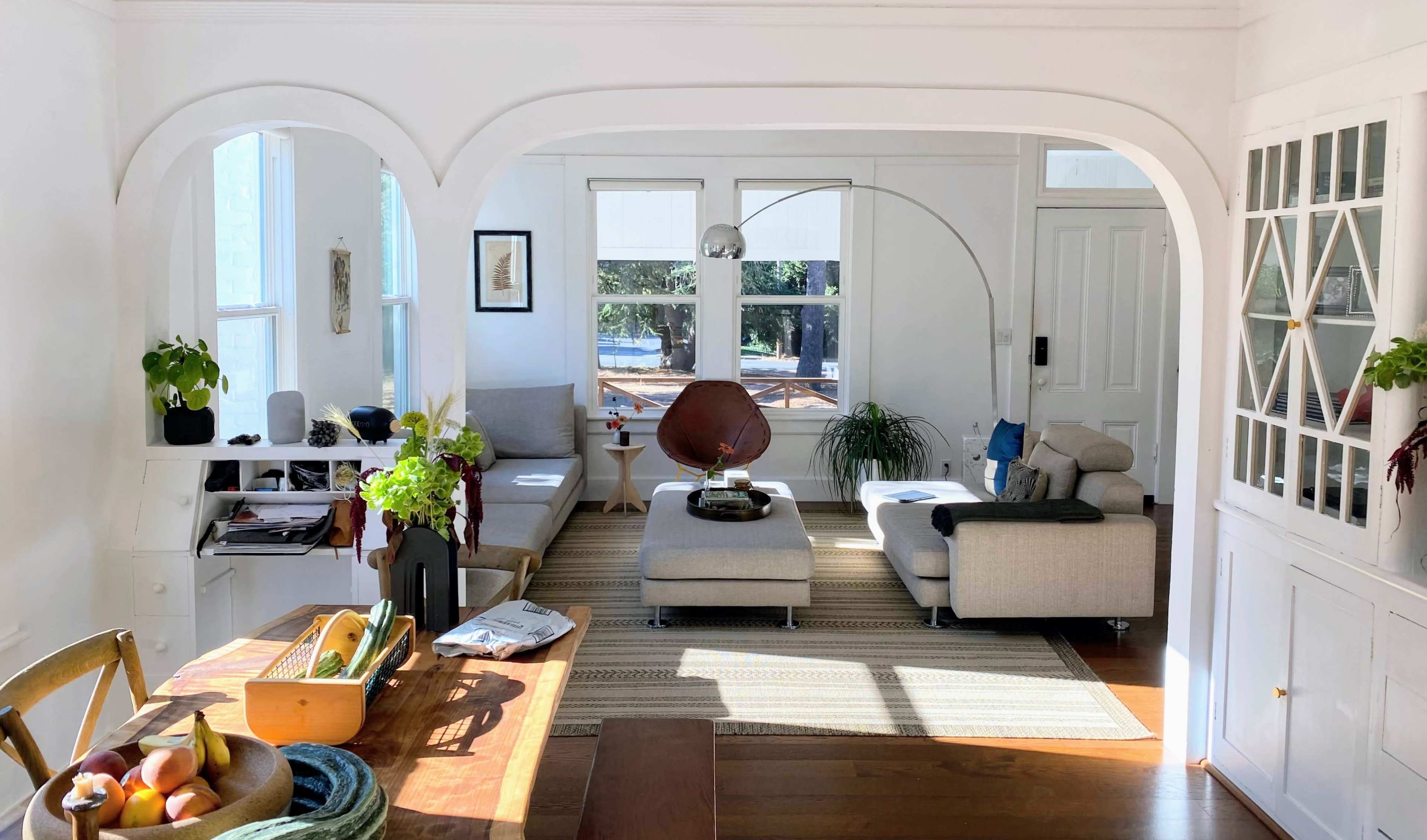 A bright, modern living room features a curved archway, a large window, a minimalist gray sectional sofa, and a wooden dining table with plants and decor.