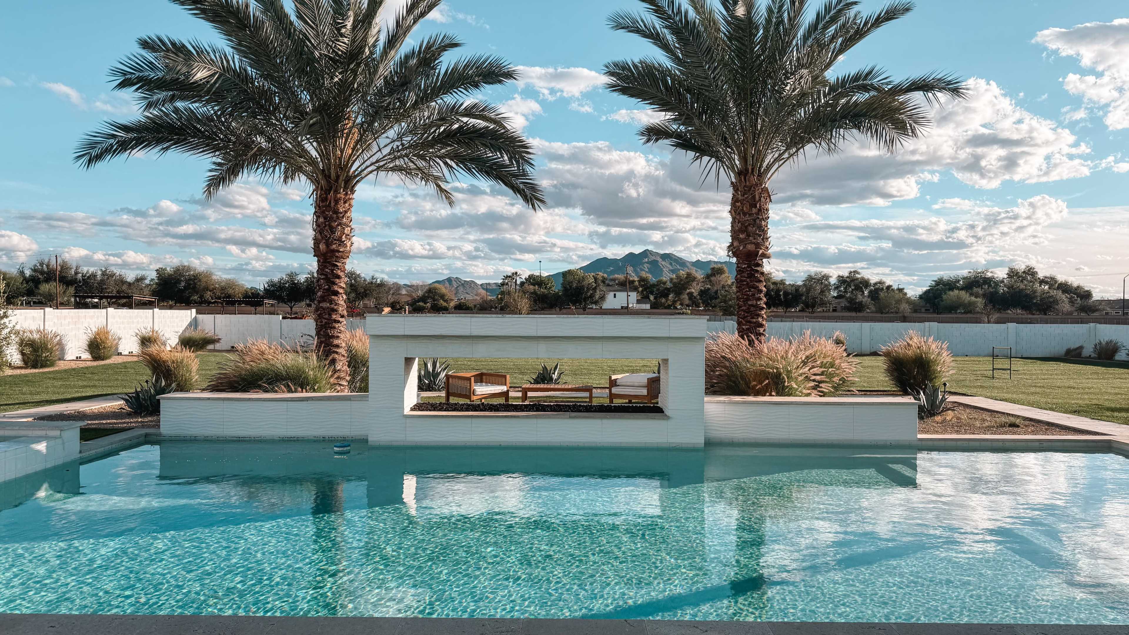 A tranquil scene features a swimming pool surrounded by palm trees, with a seating area framed by a white wall and distant mountains under a partly cloudy sky.