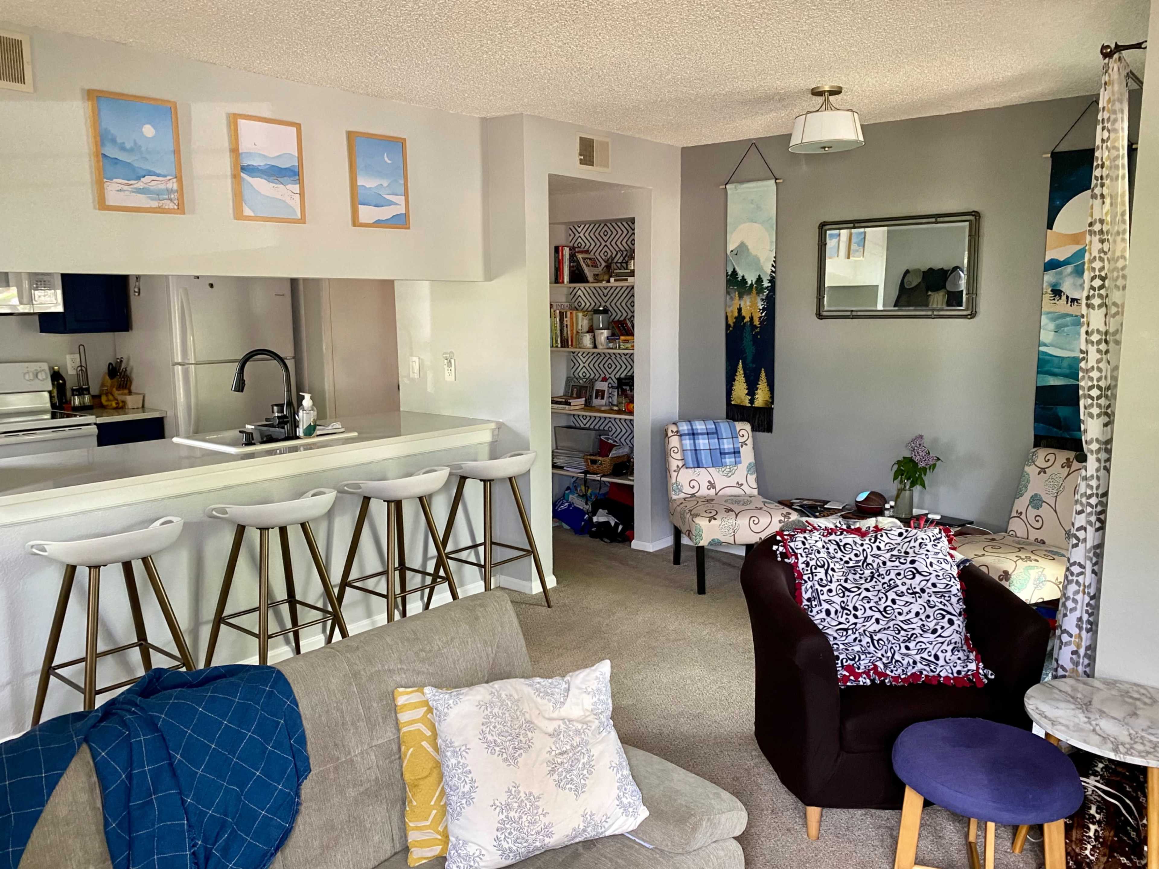 The image shows a cozy living room with a kitchen area, featuring bar stools, framed landscape art on the walls, and various seating arrangements.
