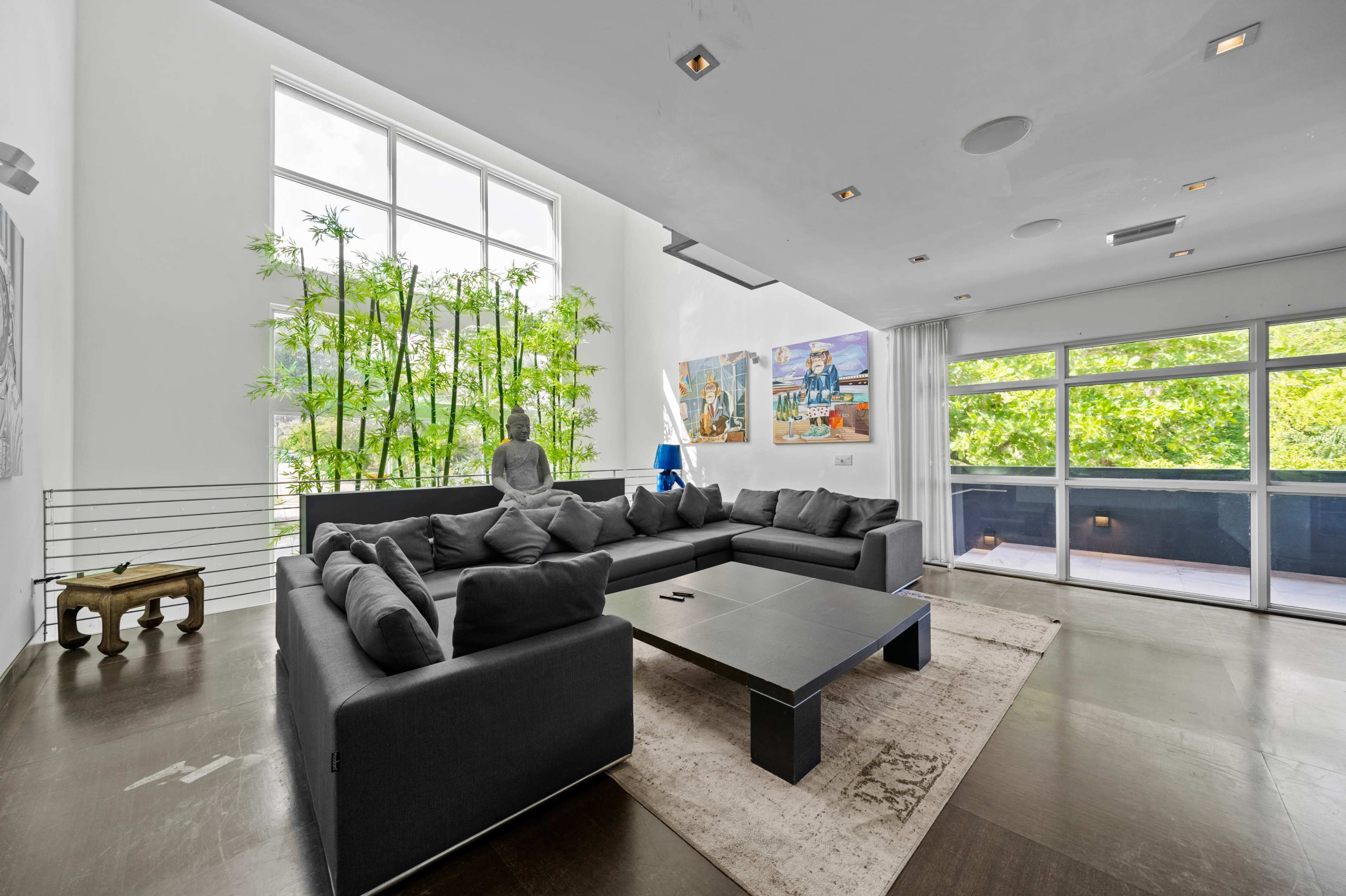 A modern living room features a large sectional sofa, a minimalistic coffee table, artwork on the walls, and a tall window with greenery visible outside.