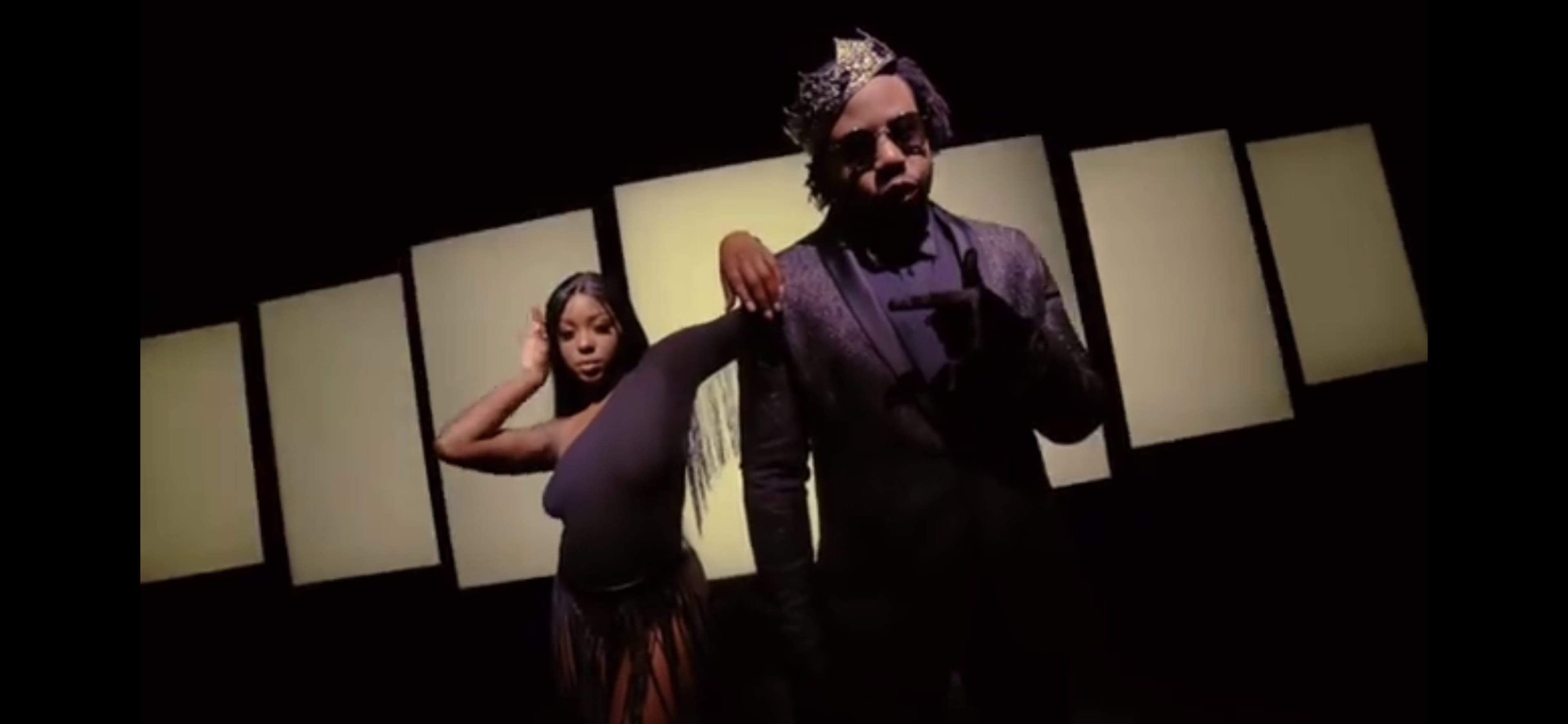 A man wearing sunglasses and a crown poses with his arm extended while a woman in a black outfit stands behind him with one arm raised.