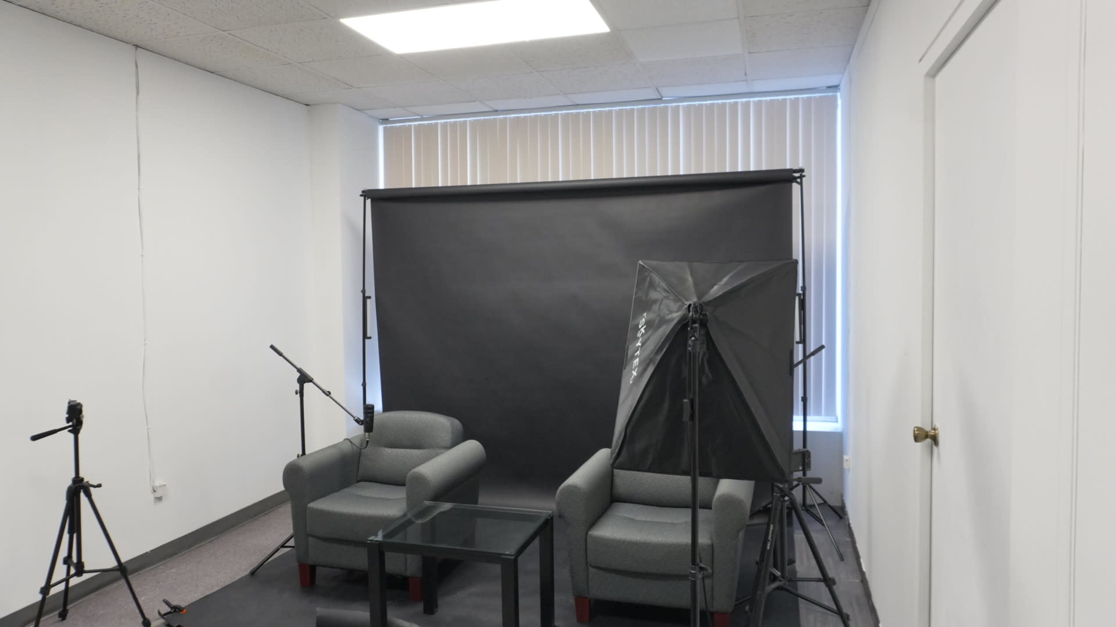 The image shows a small studio setup with two gray armchairs, a glass table, a black backdrop, and lighting equipment.
