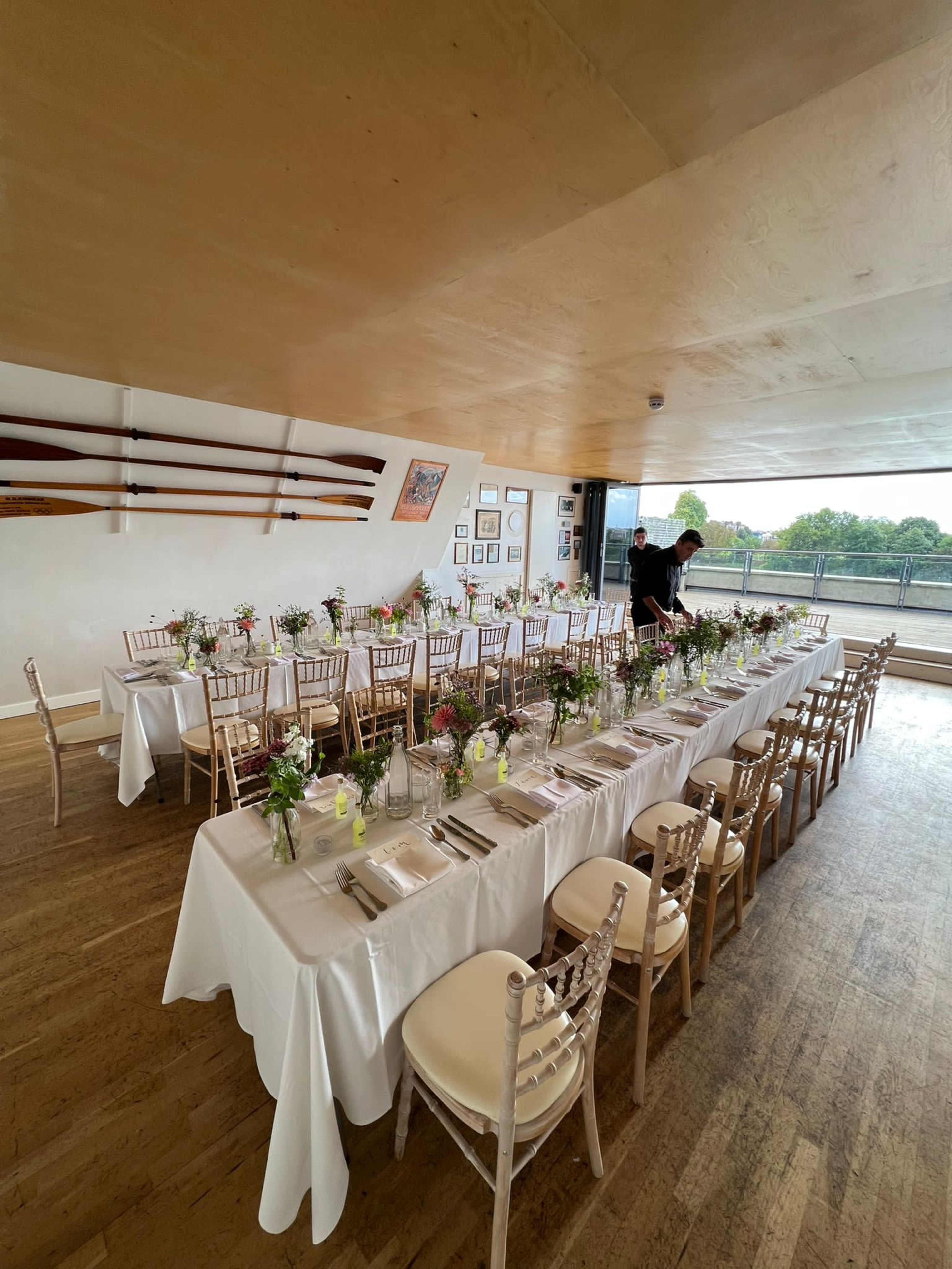 A long table set with white tablecloths and floral centerpieces is arranged in a dining room with large windows overlooking a view.
