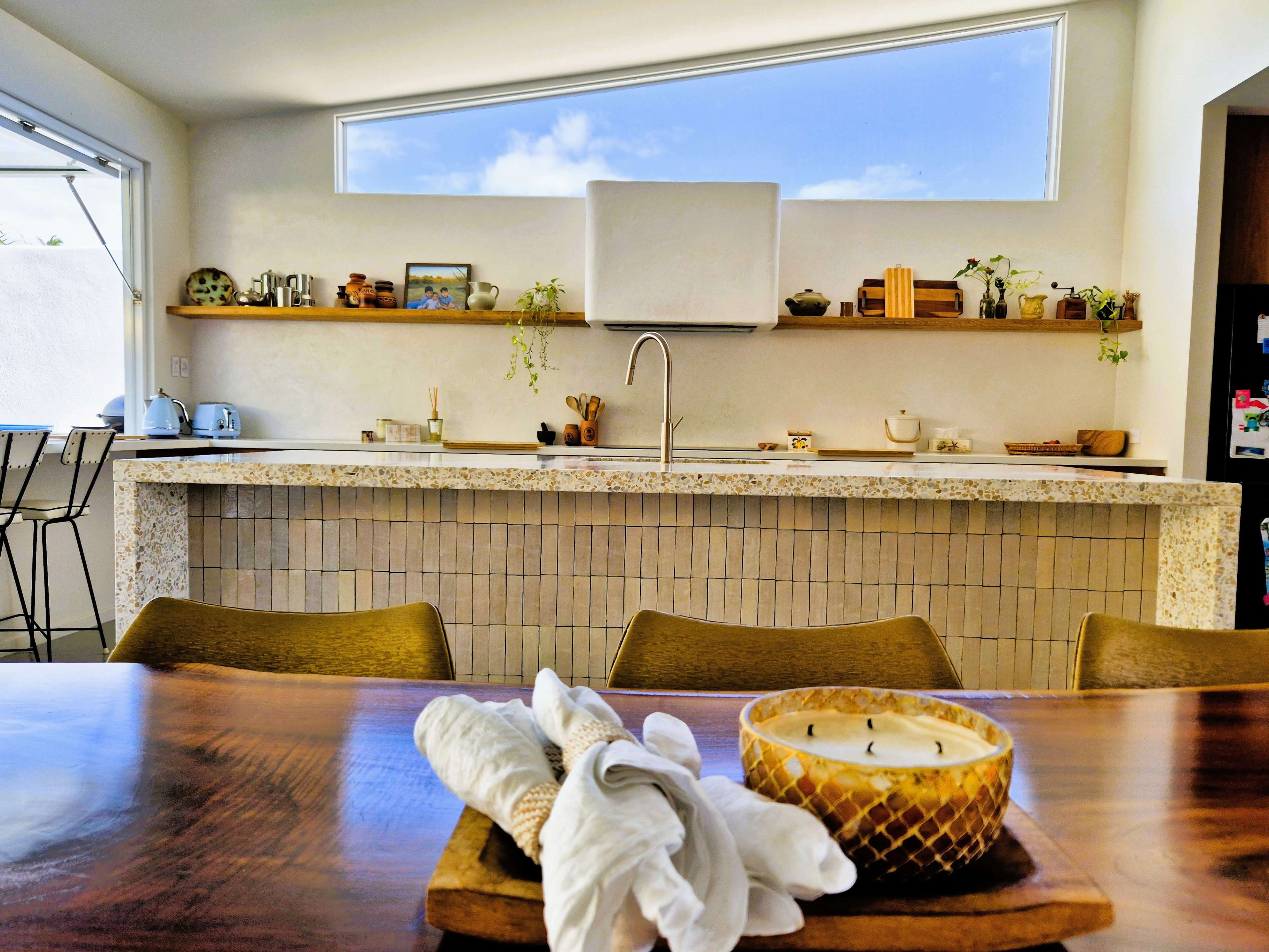 The image shows a modern kitchen with a stone countertop, wooden shelves holding various kitchen items, and a dining table set with a candle and cloth napkins.