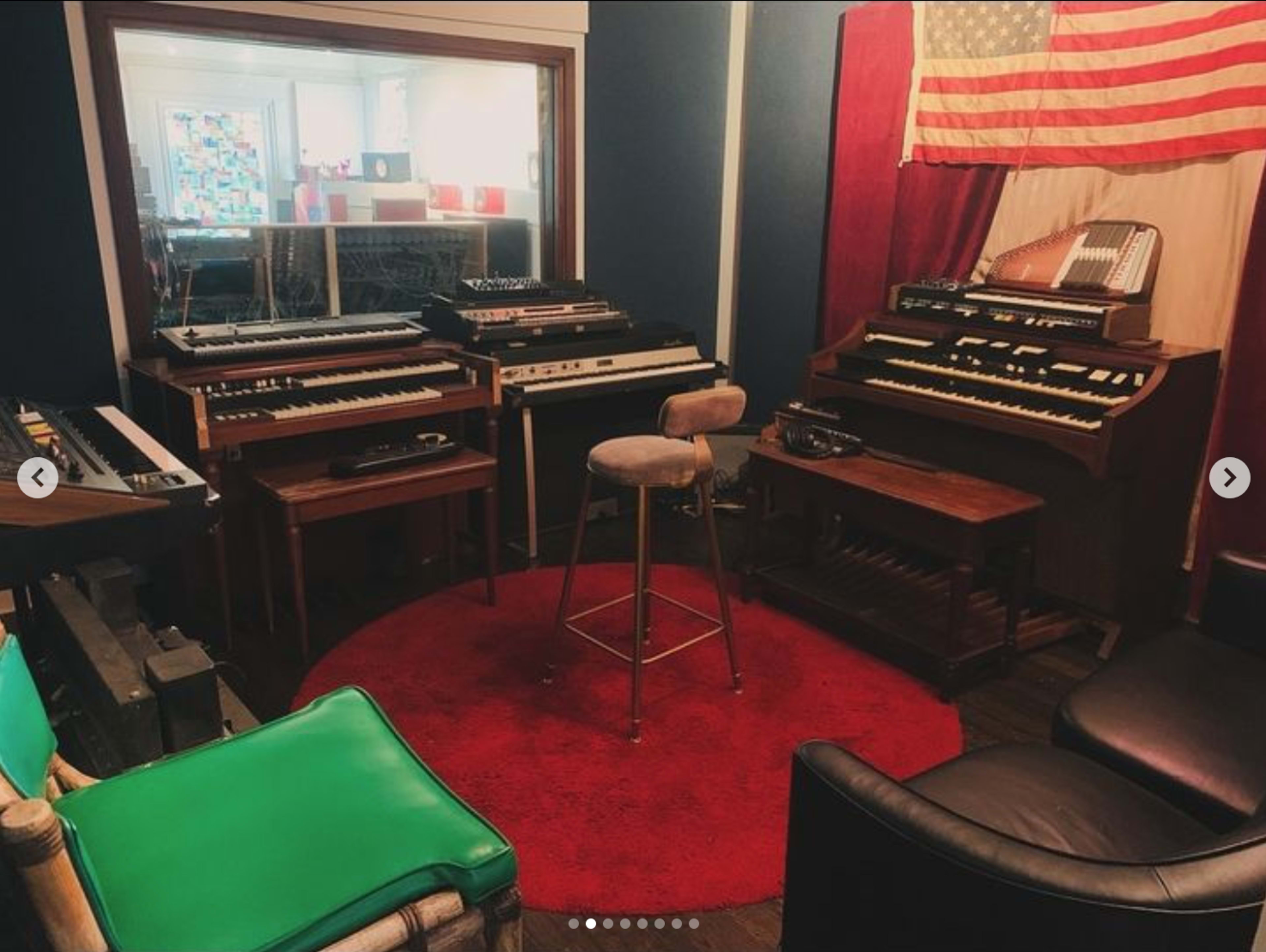 The image showcases a cozy music studio featuring various vintage keyboards, a stool in the center, and a backdrop with an American flag.