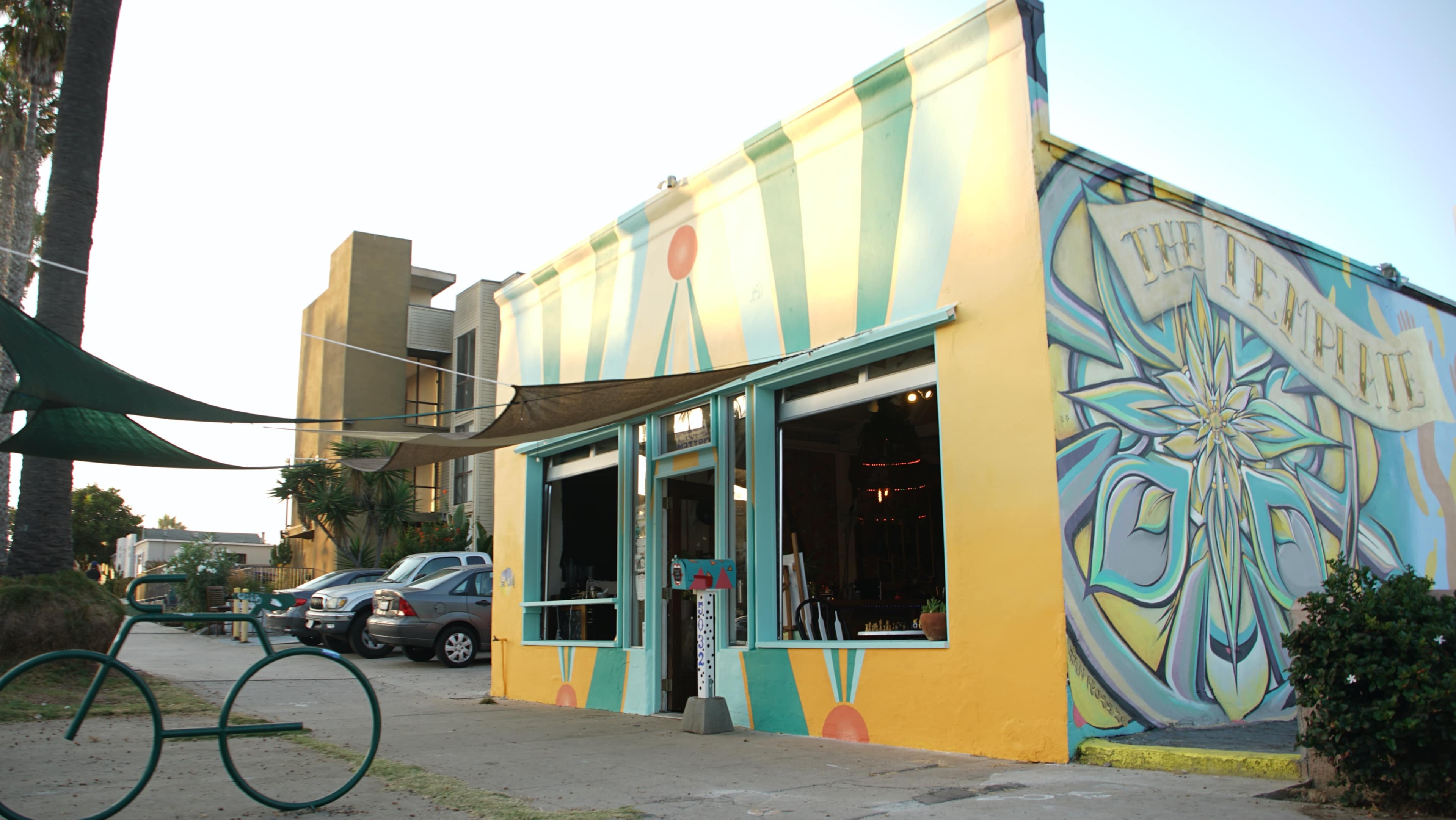 The image shows a brightly painted building with a colorful mural on its exterior, located in a street setting with vehicles parked nearby.