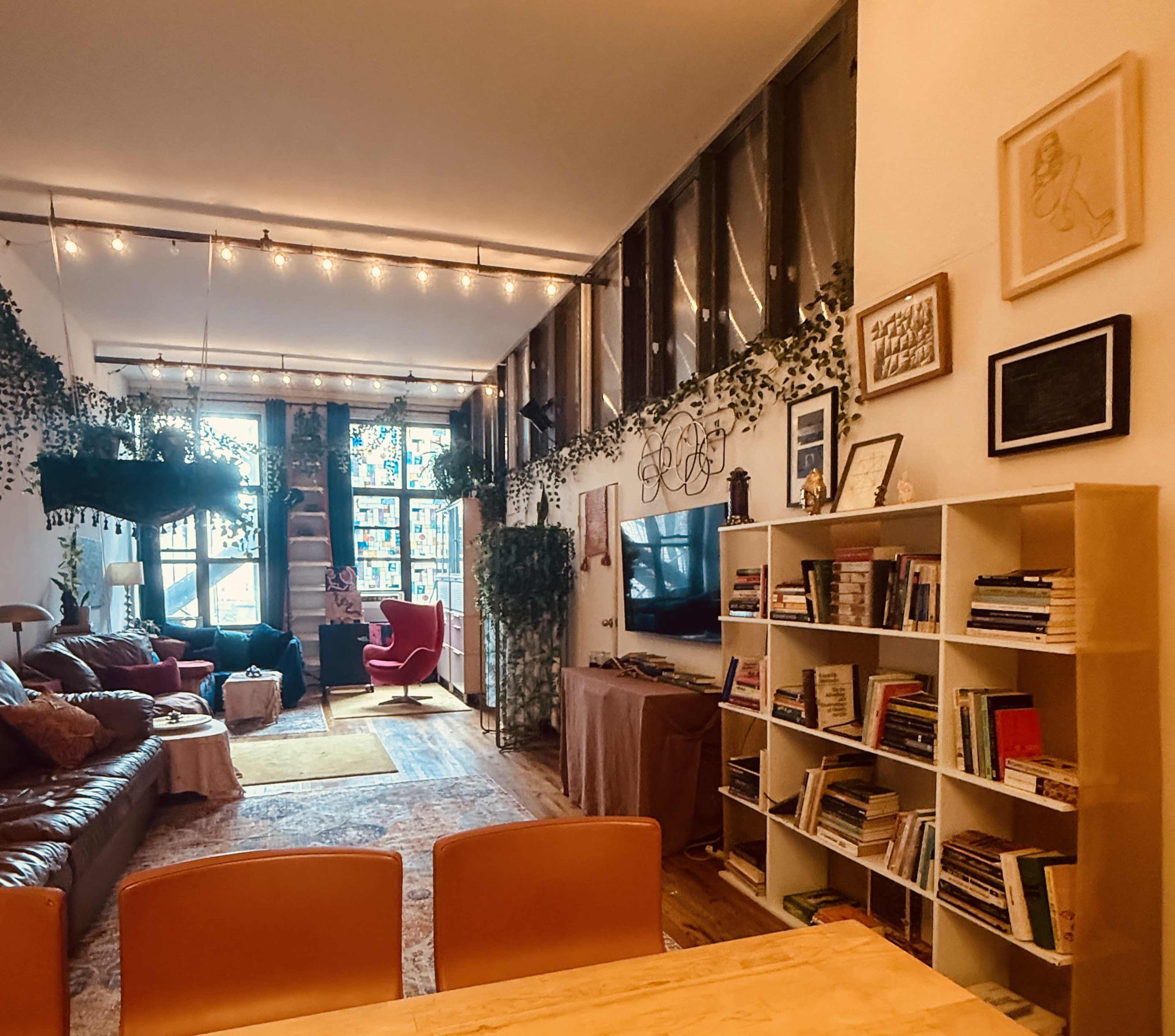 The image shows a cozy living space with a mix of seating options, shelves filled with books, and greenery adorned with string lights.