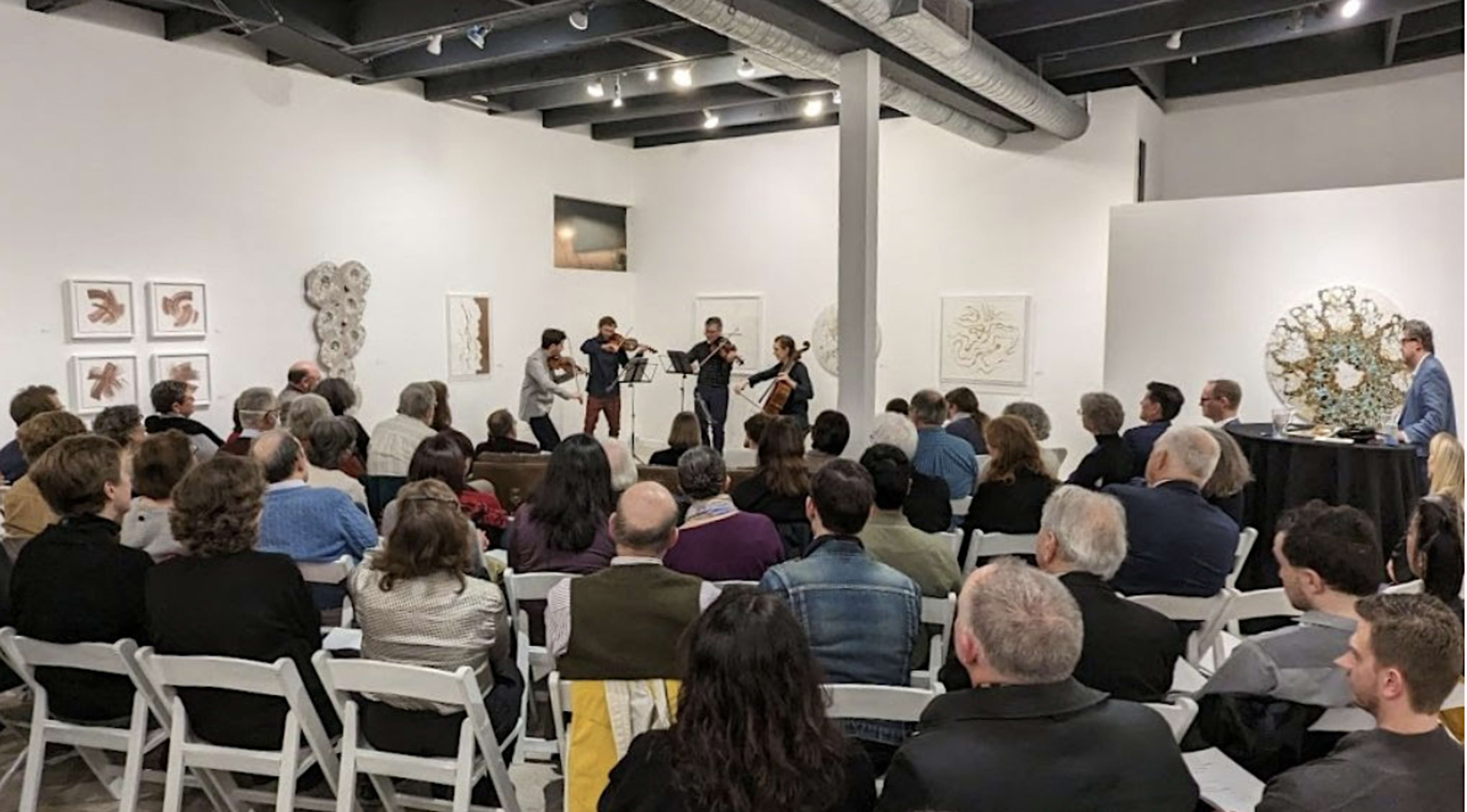 A small ensemble performs in a gallery setting filled with an audience seated on folding chairs.