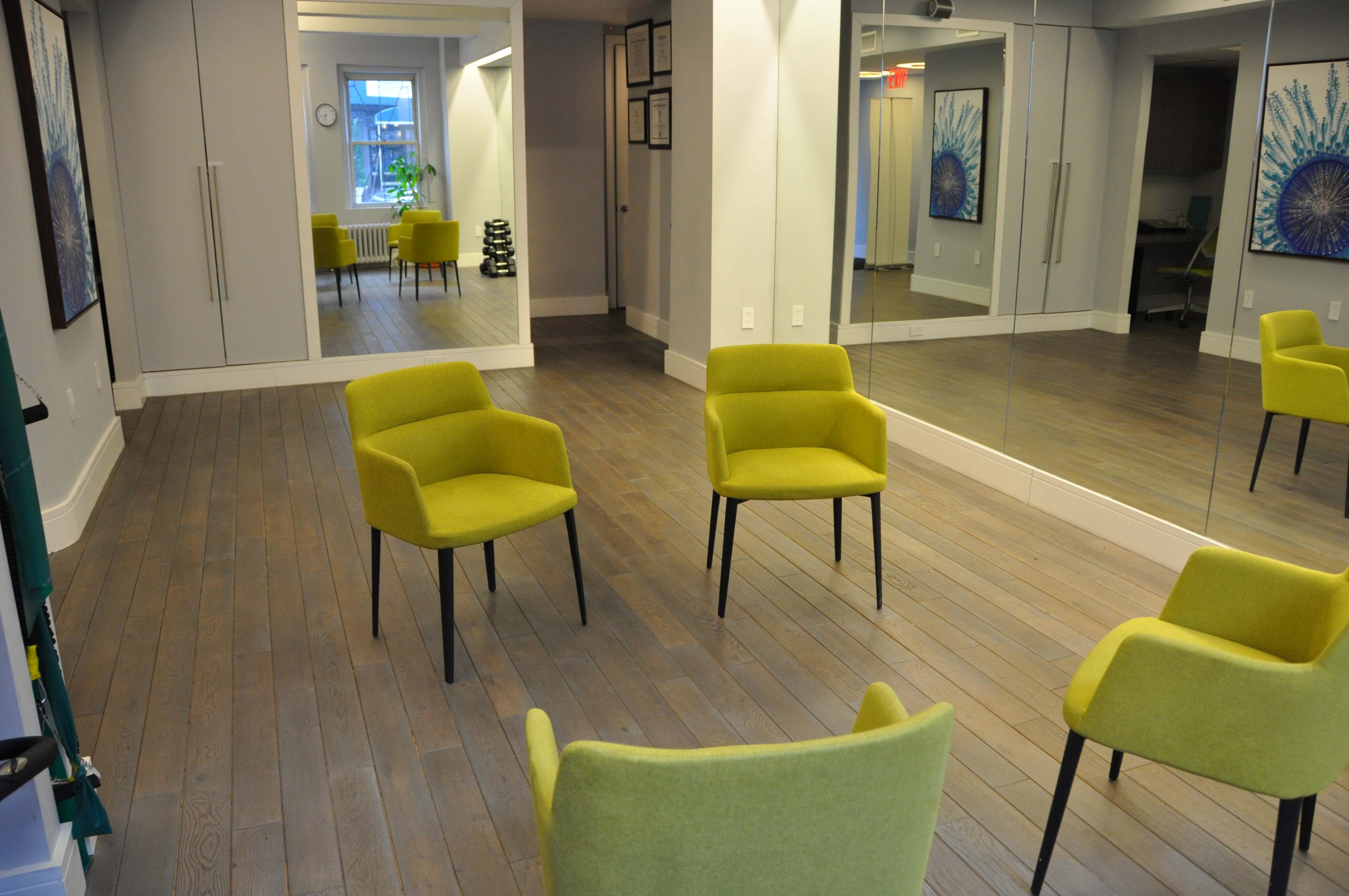 A spacious, well-lit room features four yellow chairs arranged in a circular formation on a wooden floor, with mirrors and artwork lining the walls.