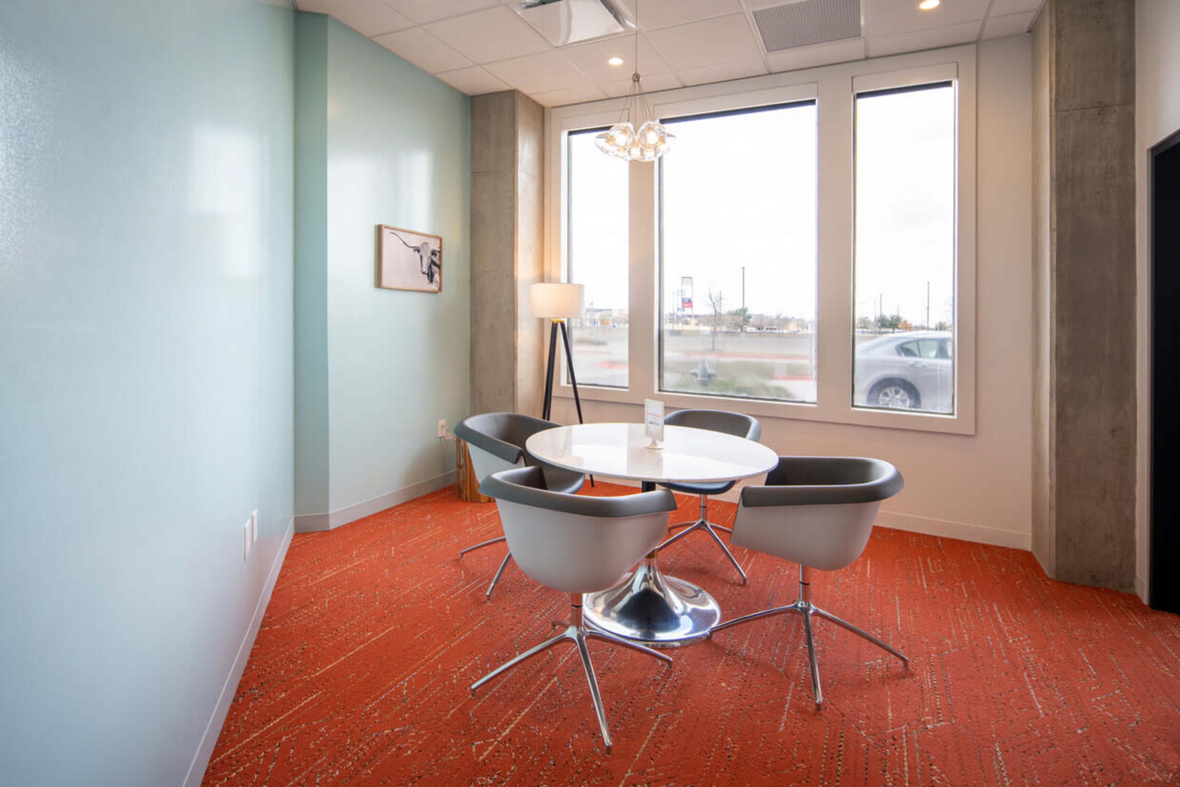 A small meeting room features a round table surrounded by four modern chairs, with large windows providing natural light and a view of the outside.